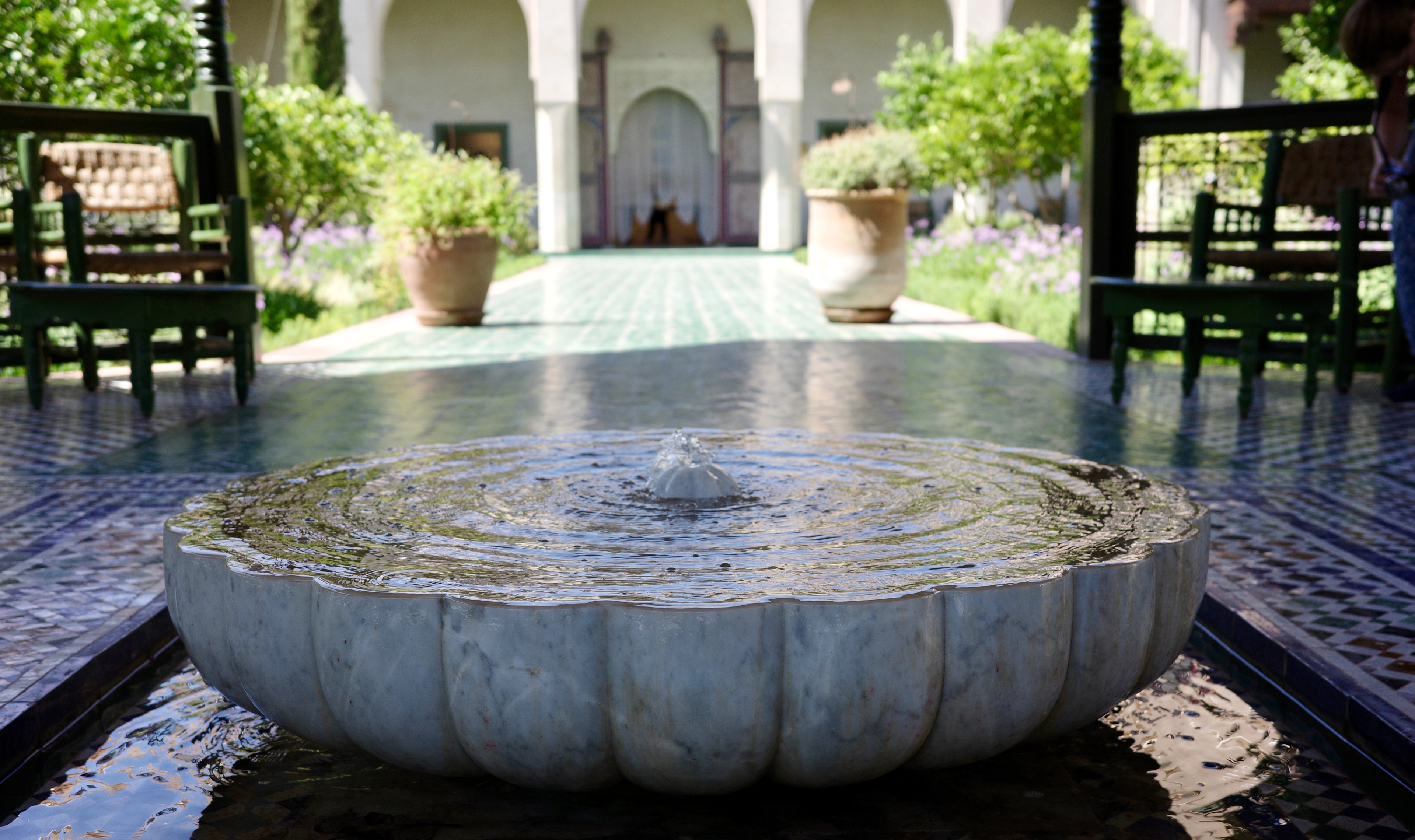 A round stone fountain stands in a shaded courtyard with plants and chairs.