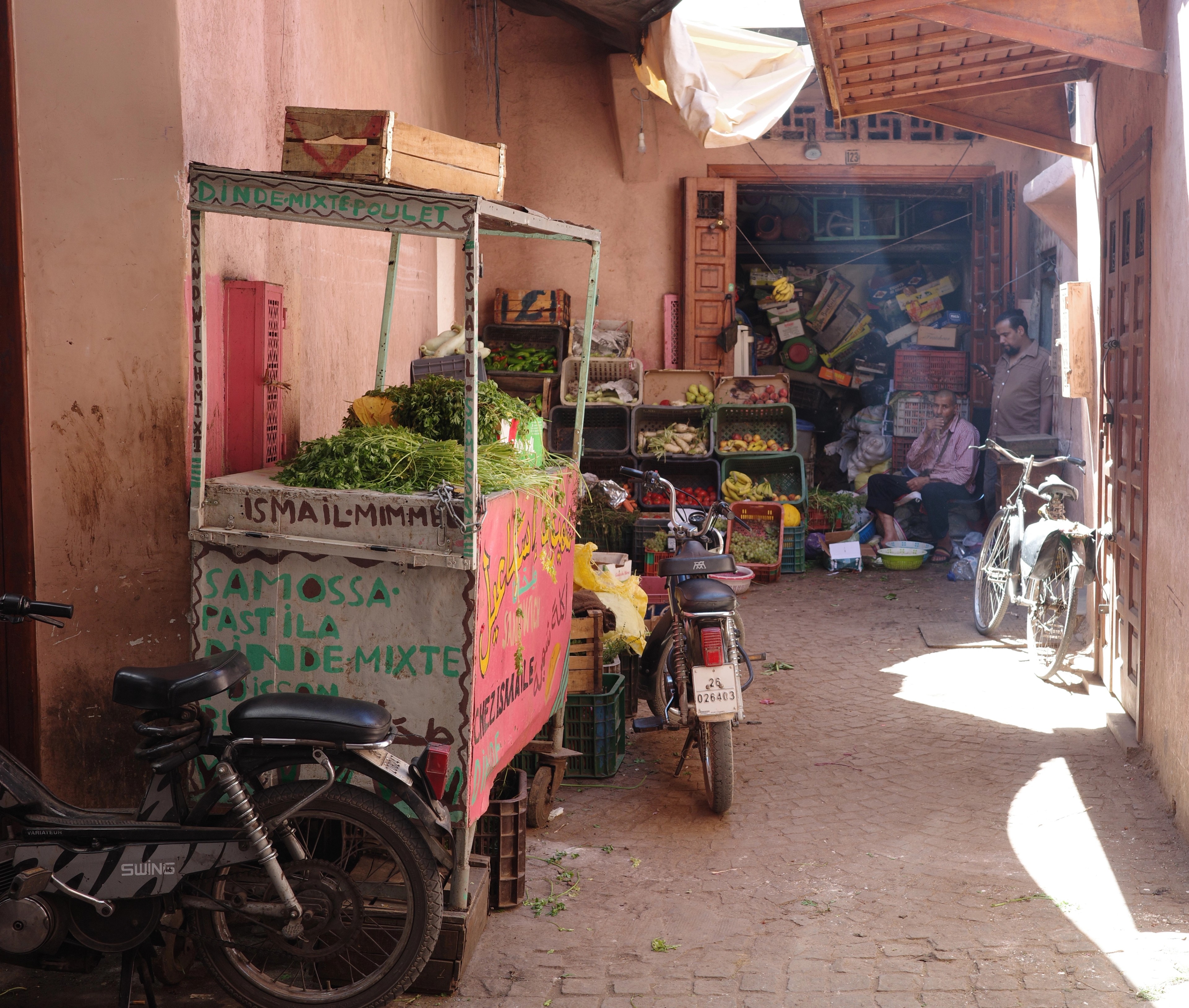 A pushcart stands in a narrow pink-walled lane lined with small shops.