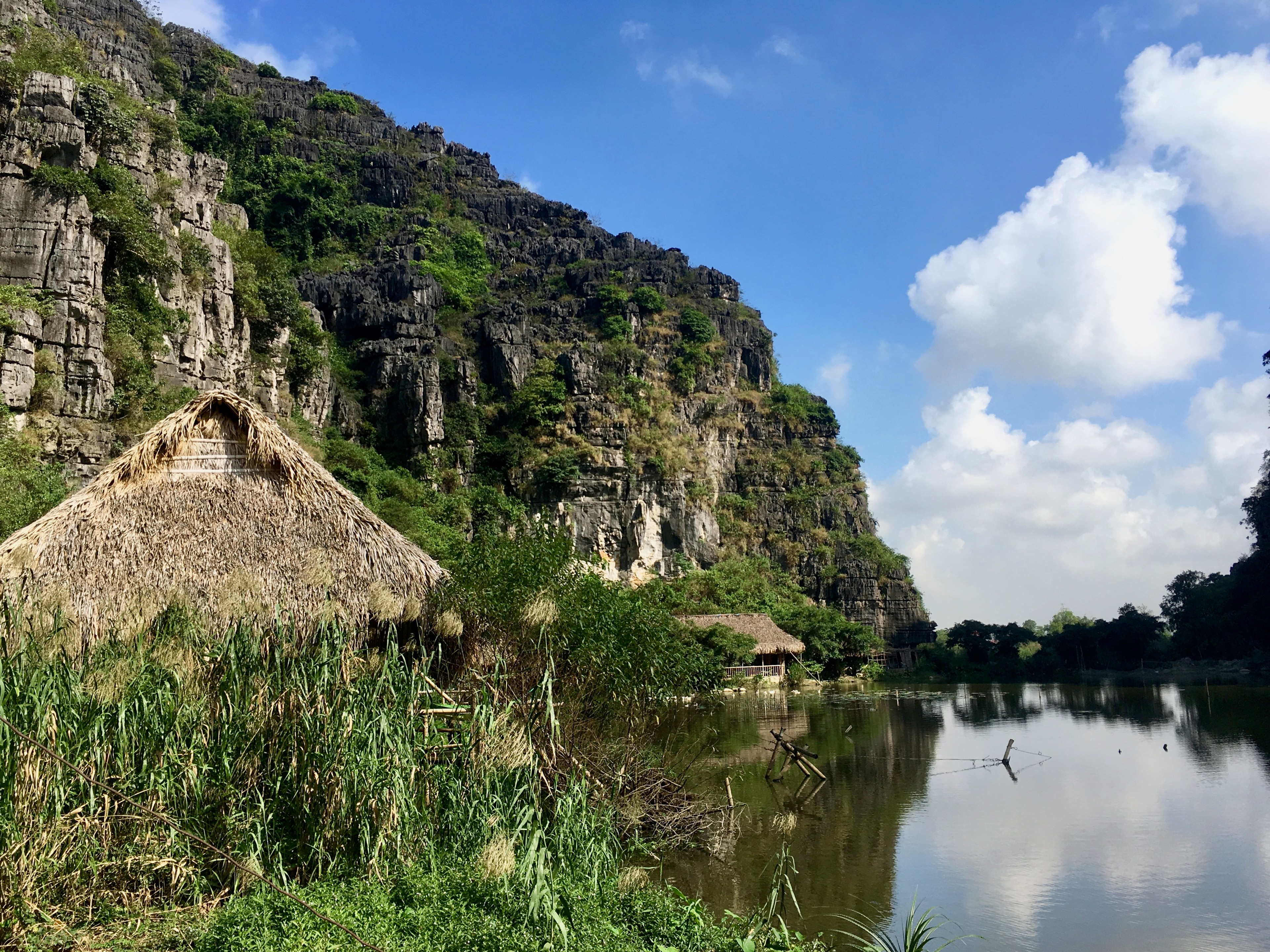 A small thatched hut sits beside a pond below a rocky green hill.