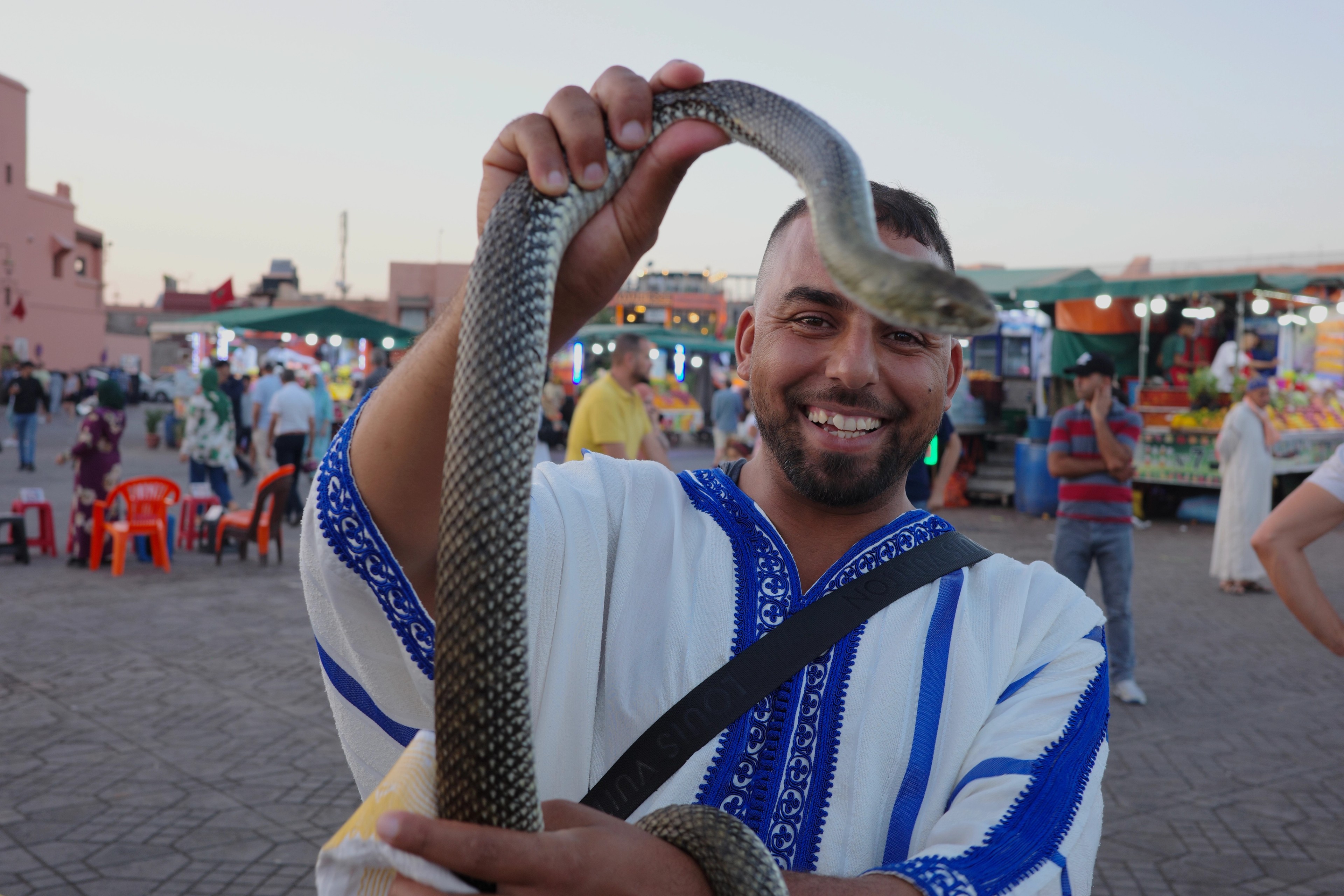 A smiling man holds a snake around his shoulders in a crowded square.