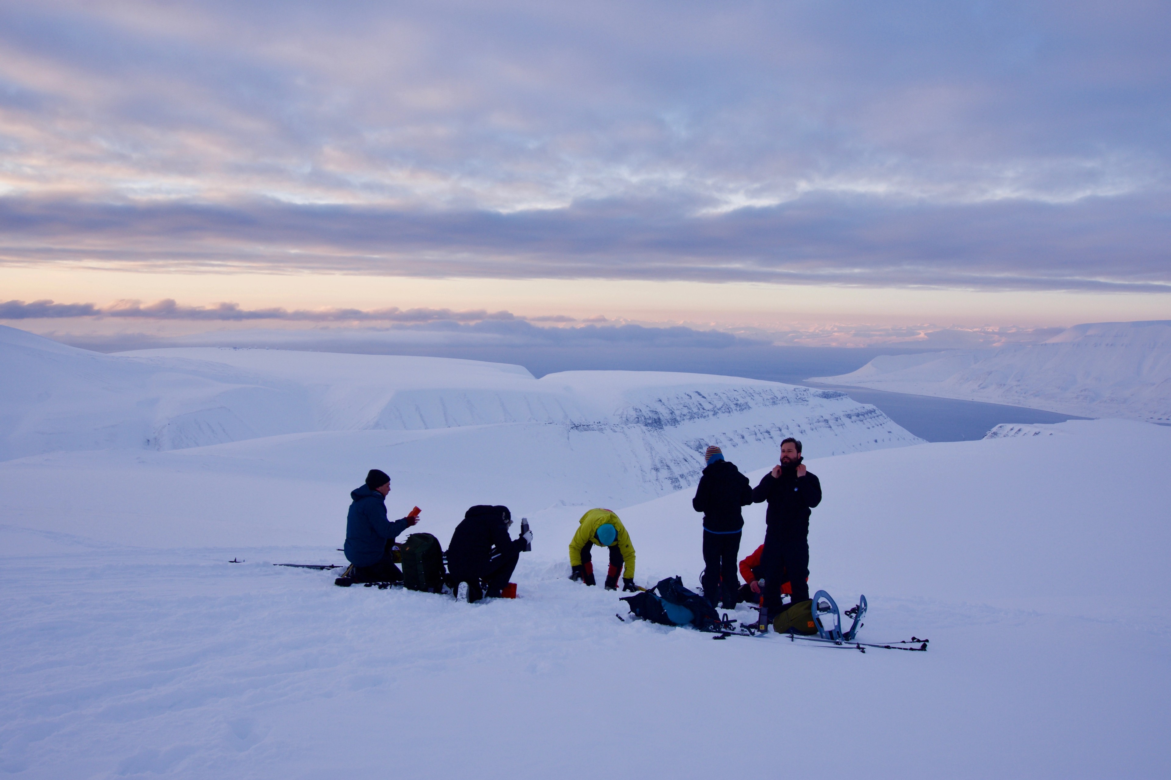 A group pauses on a snowy plain beneath a pink evening sky.