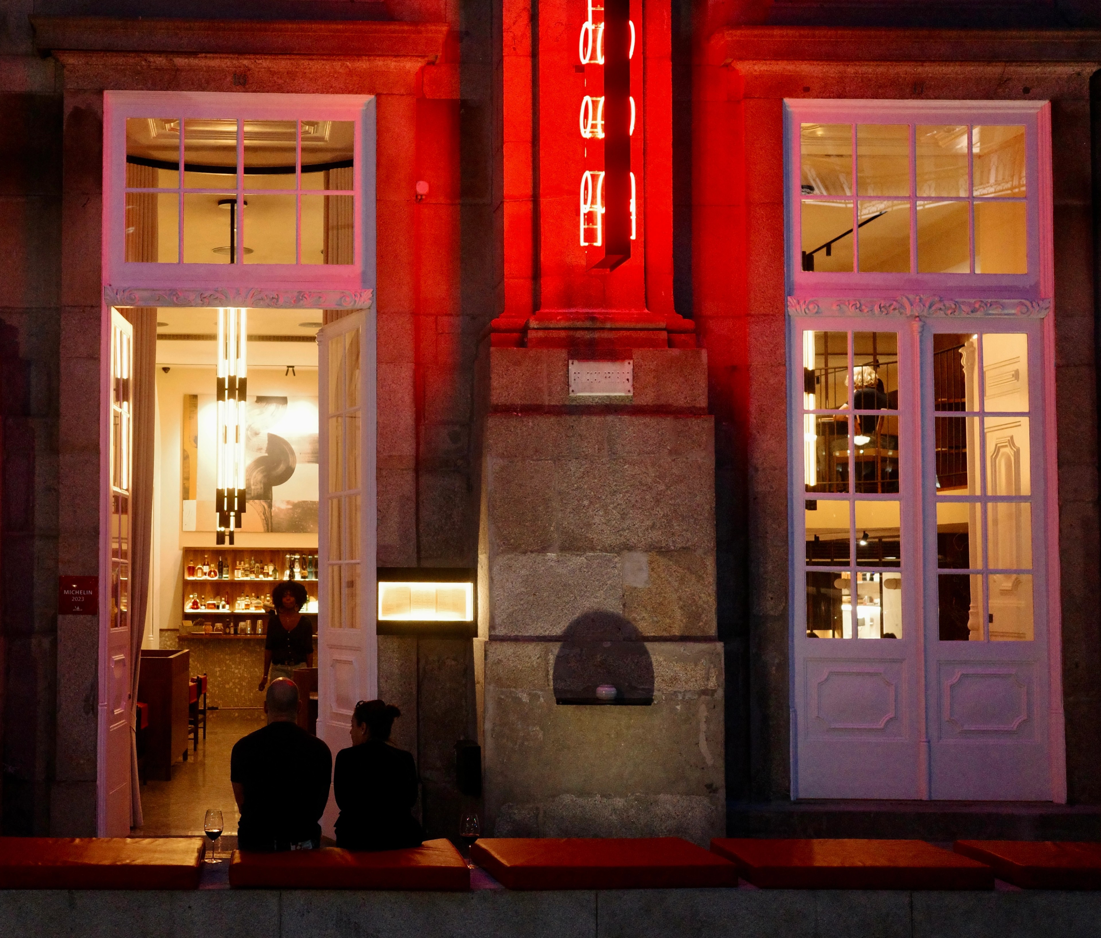 People sit outside a brightly illuminated storefront beneath a red sign.