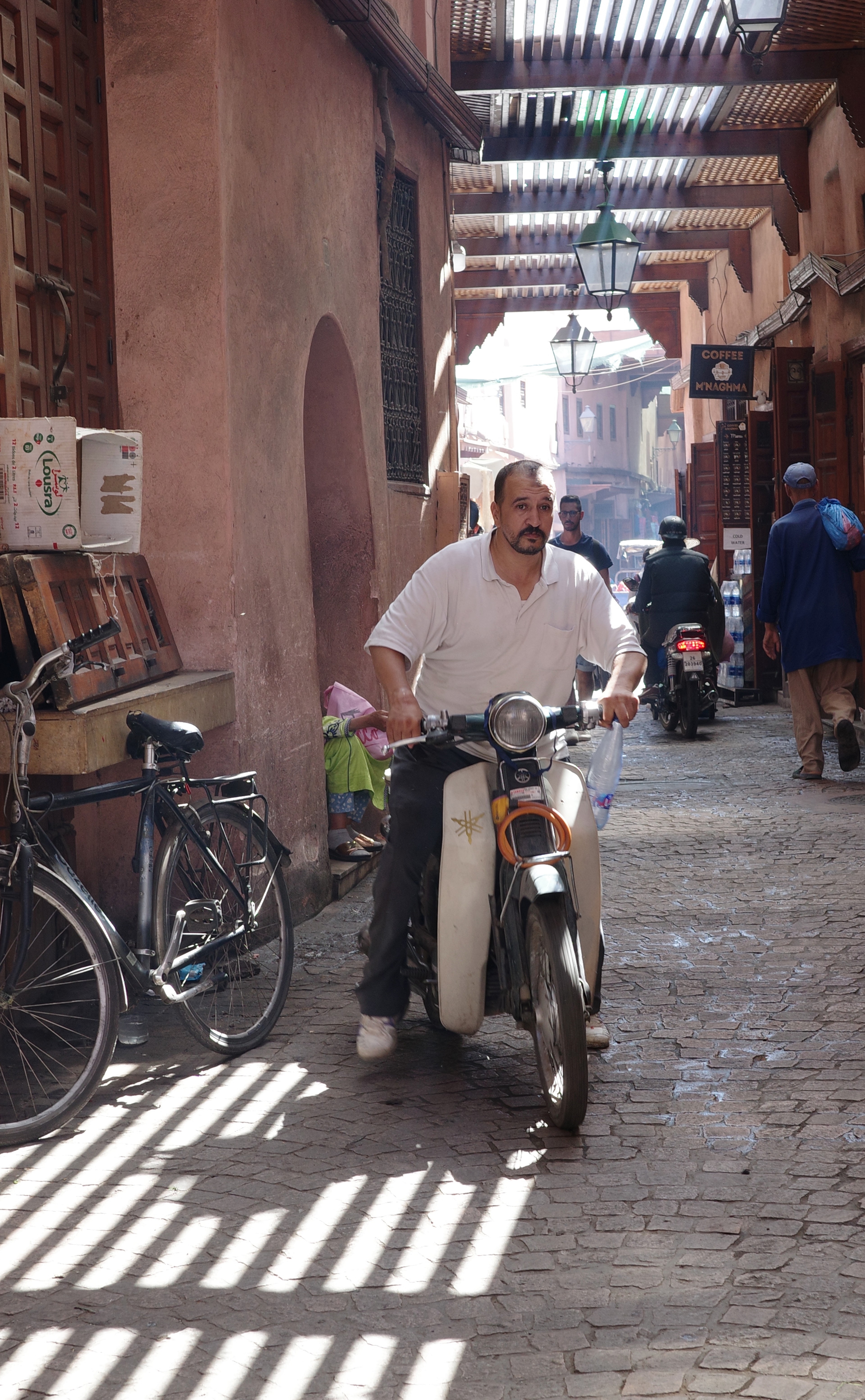 People and bicycles move through a shaded market alley.