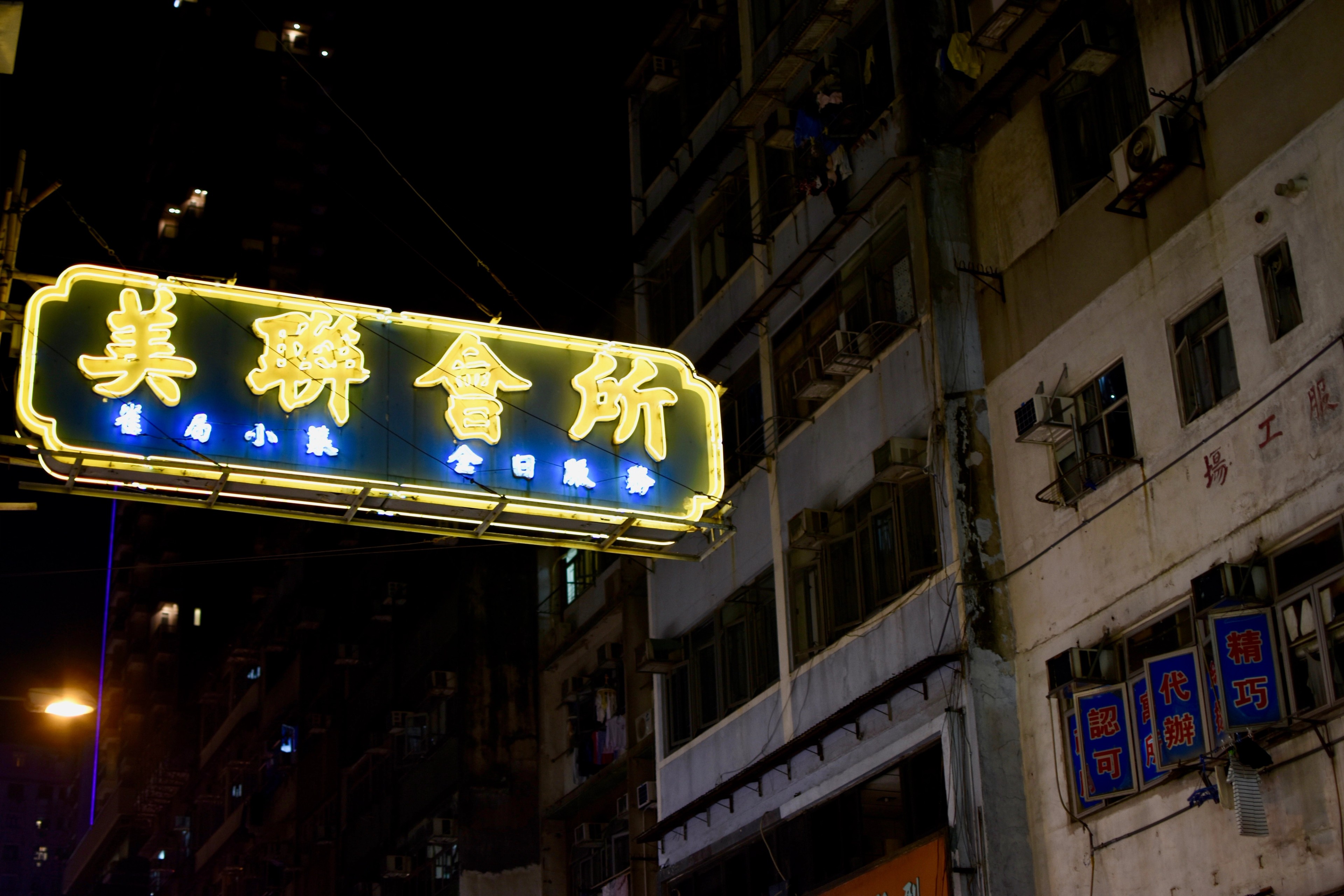 A yellow neon sign hangs above a dim street lined with apartment buildings.