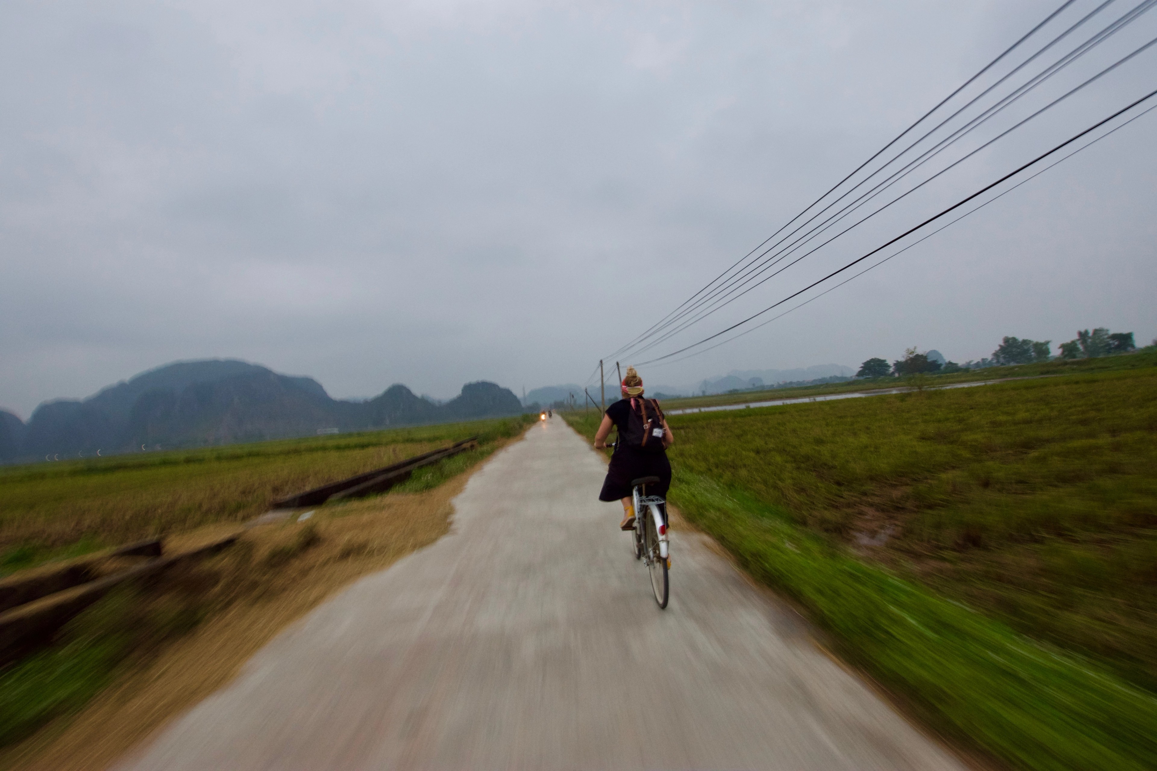 A cyclist rides along a narrow road through flat green fields beneath power lines.