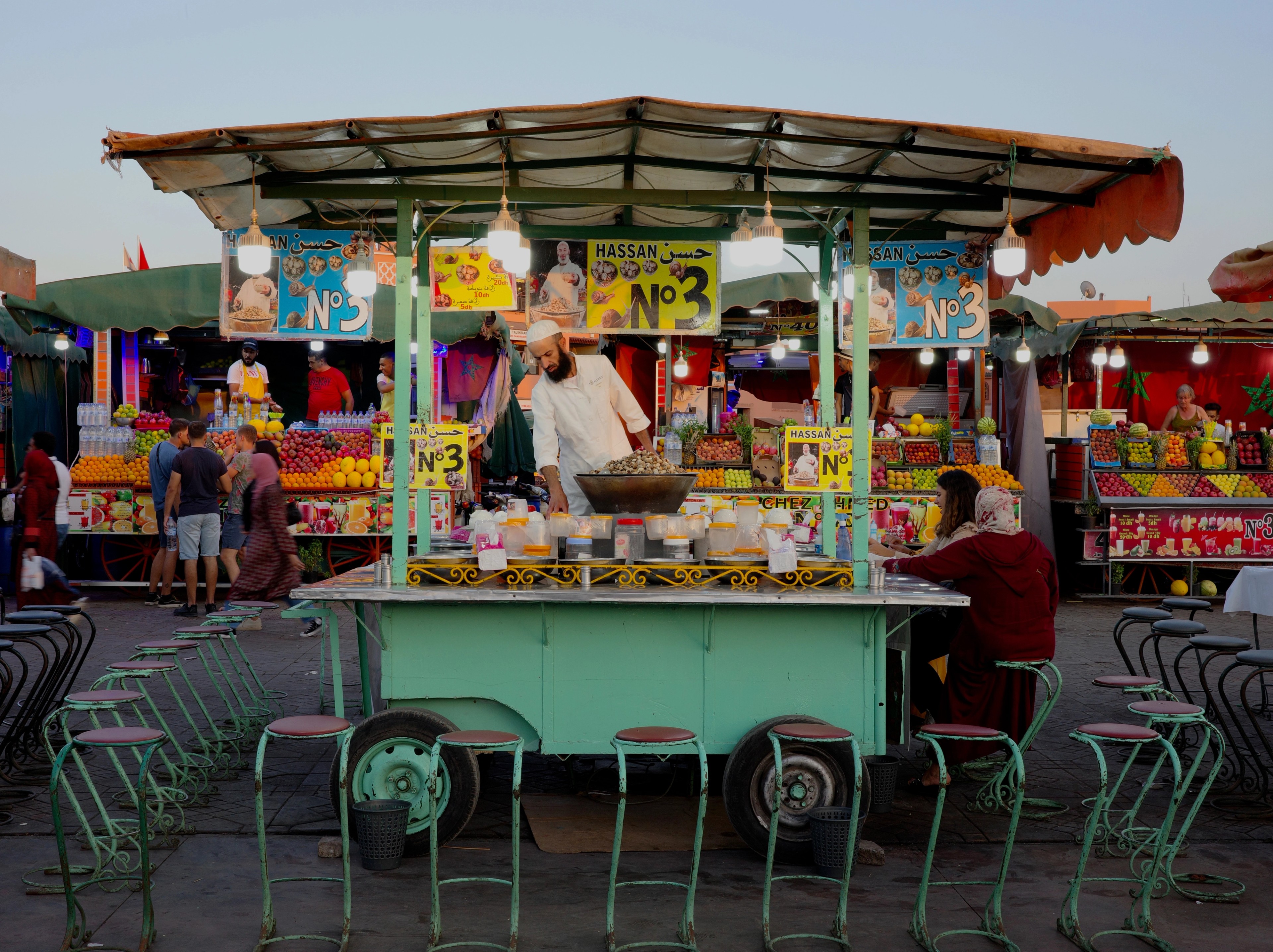 A colorful food stand with stools and hanging decorations fills a market lane.