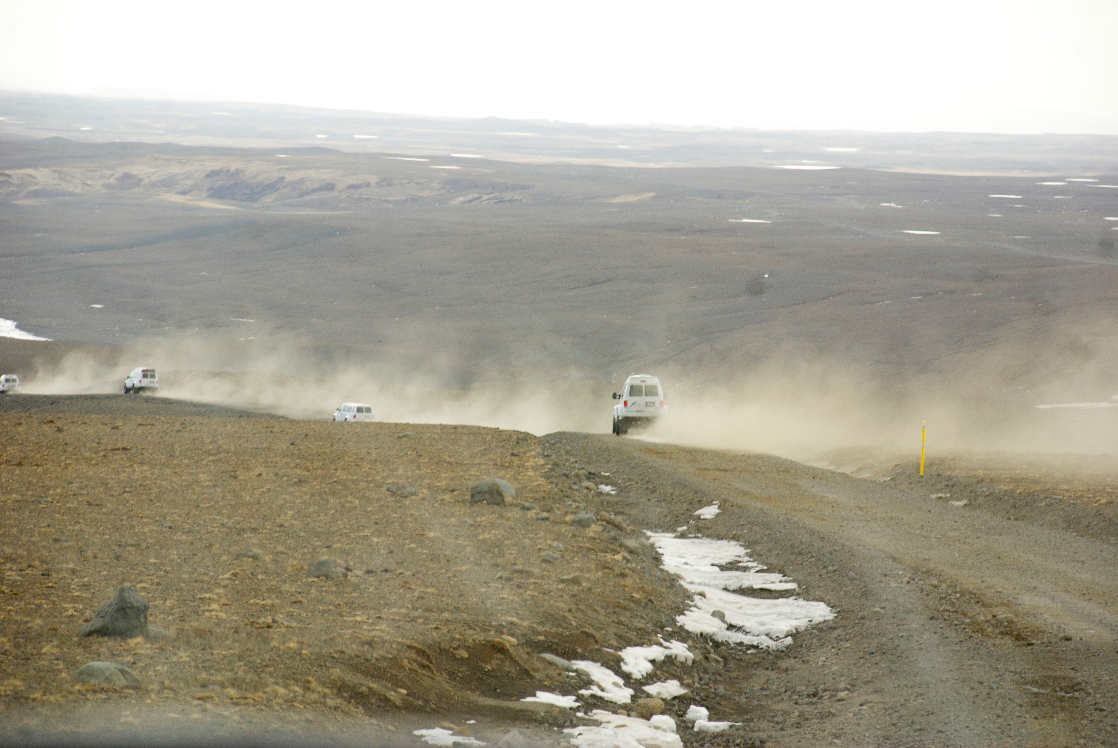 A white vehicle drives across a barren plain and raises a trail of dust.