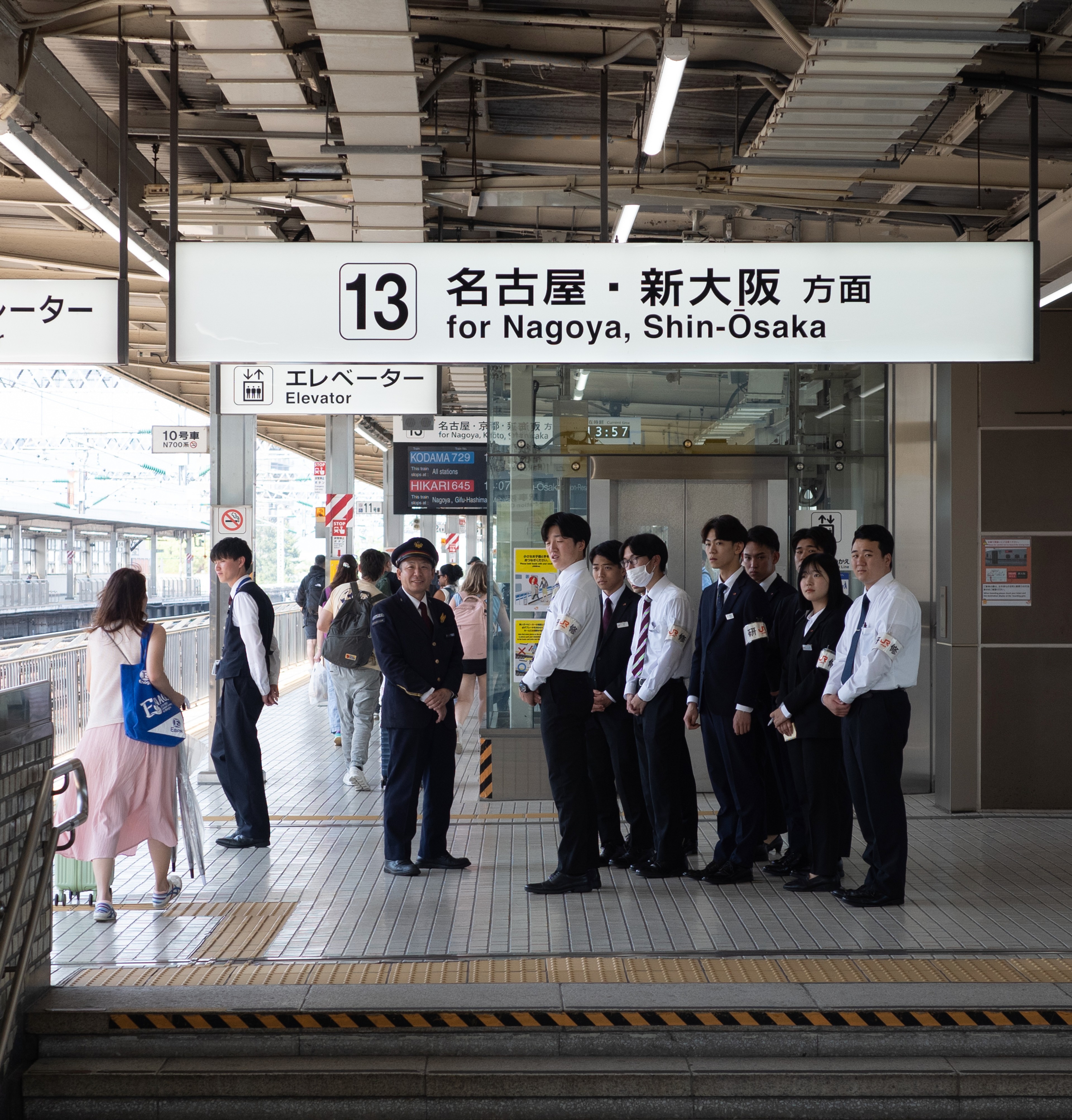 Passengers wait on a train platform beneath an overhead station sign.