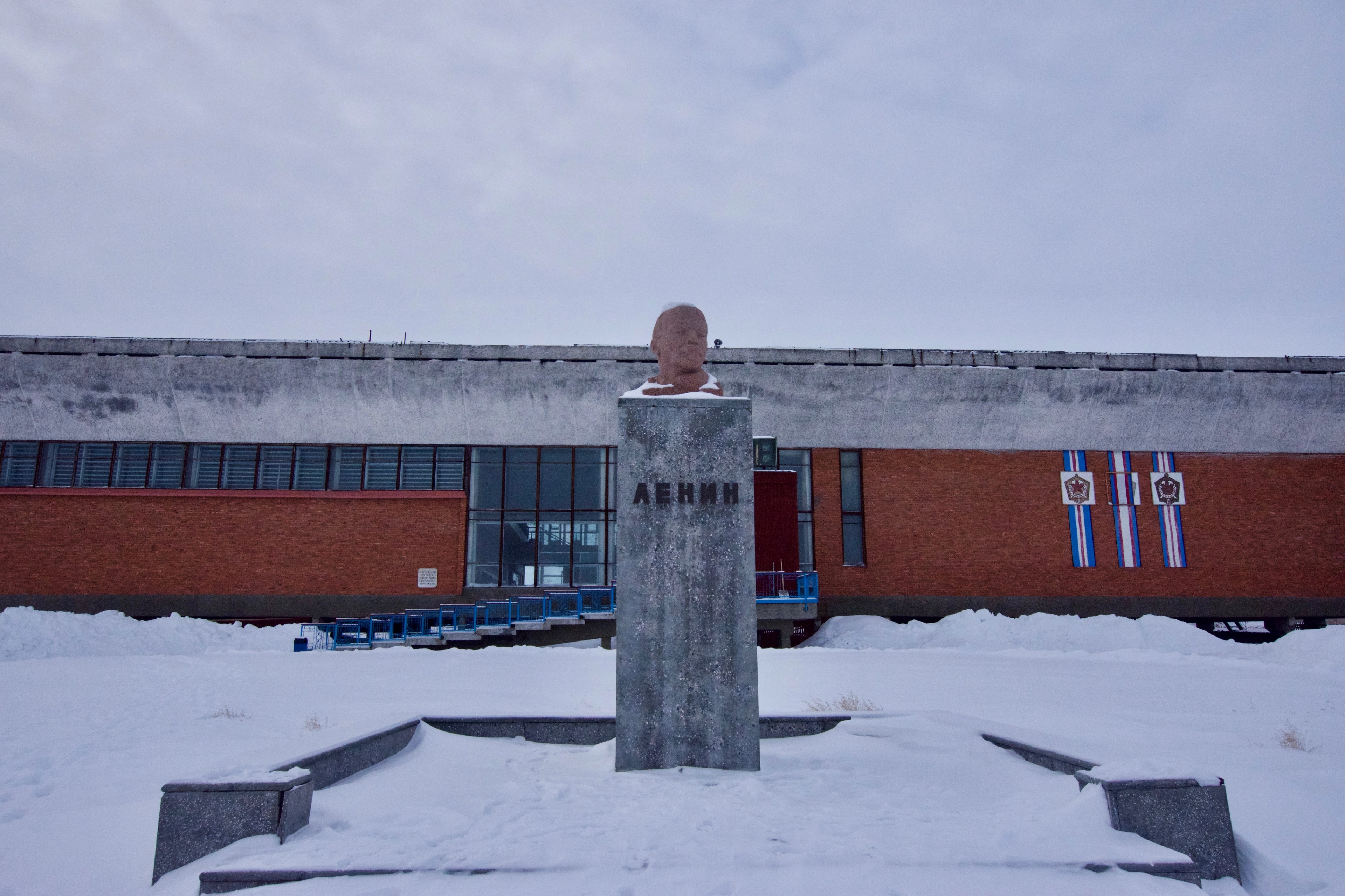 A statue stands before a long red building in the snow.