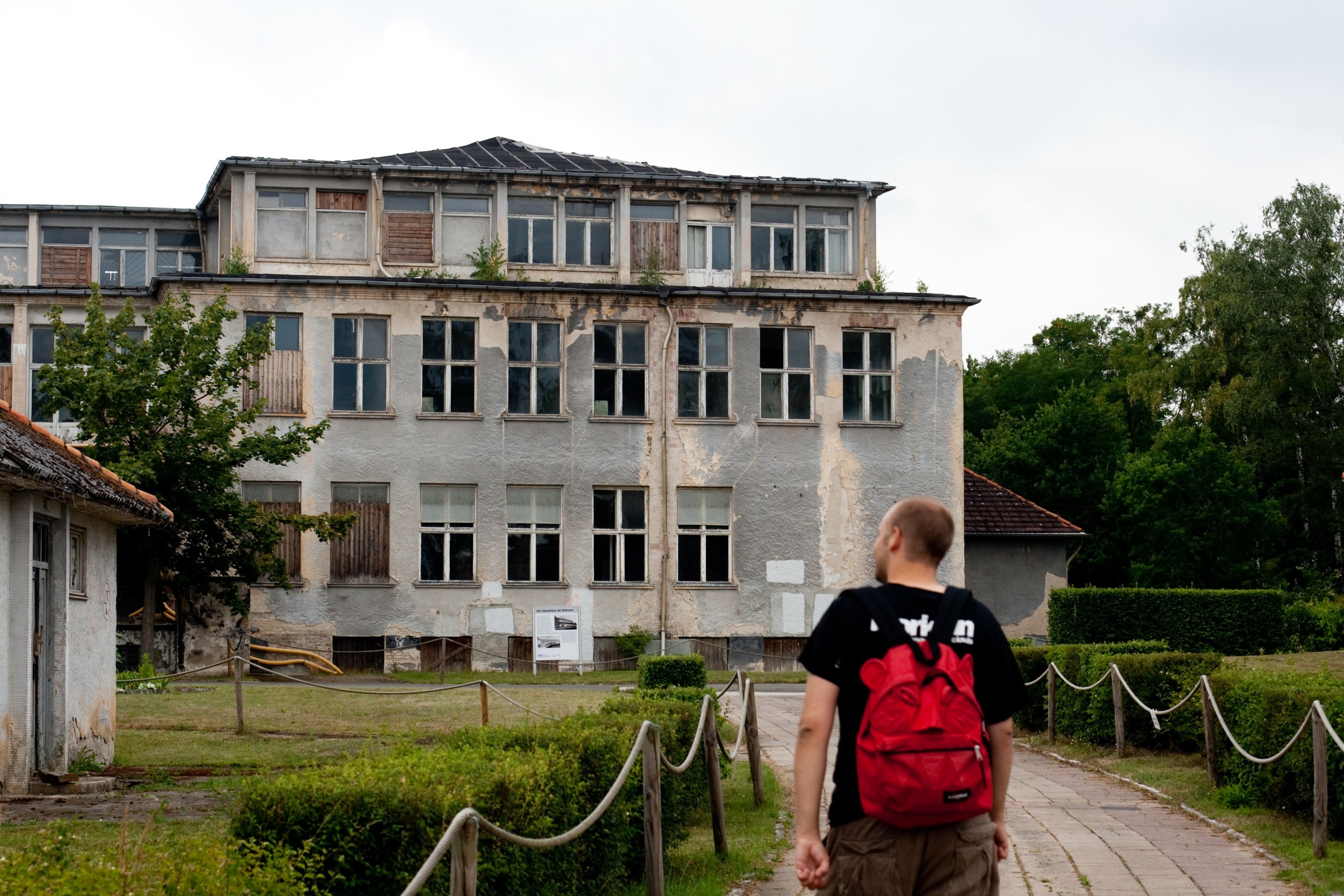 A person with a red backpack walks toward a large pale building in a green courtyard.