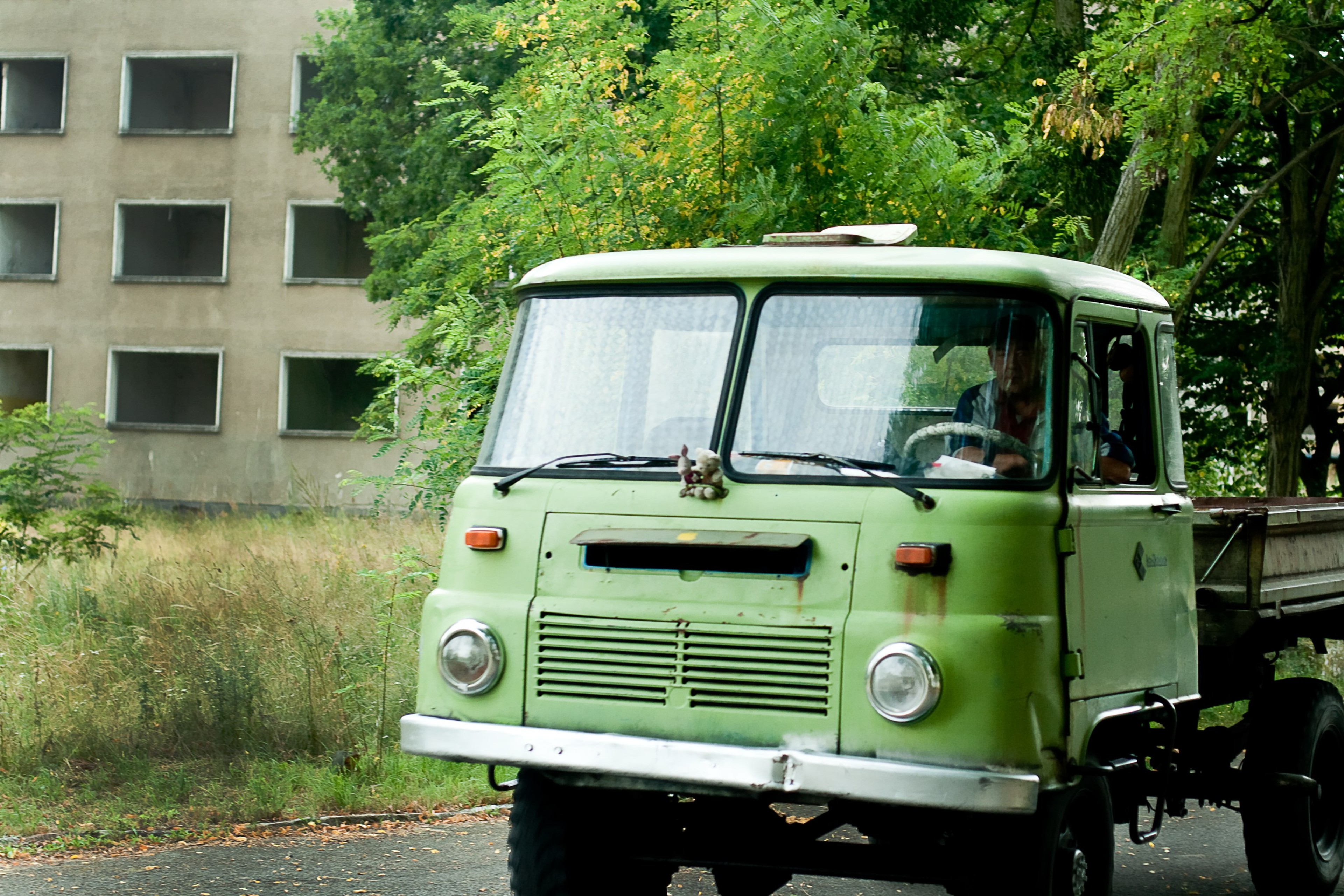 A pale green vintage van is parked beside a building and trees.