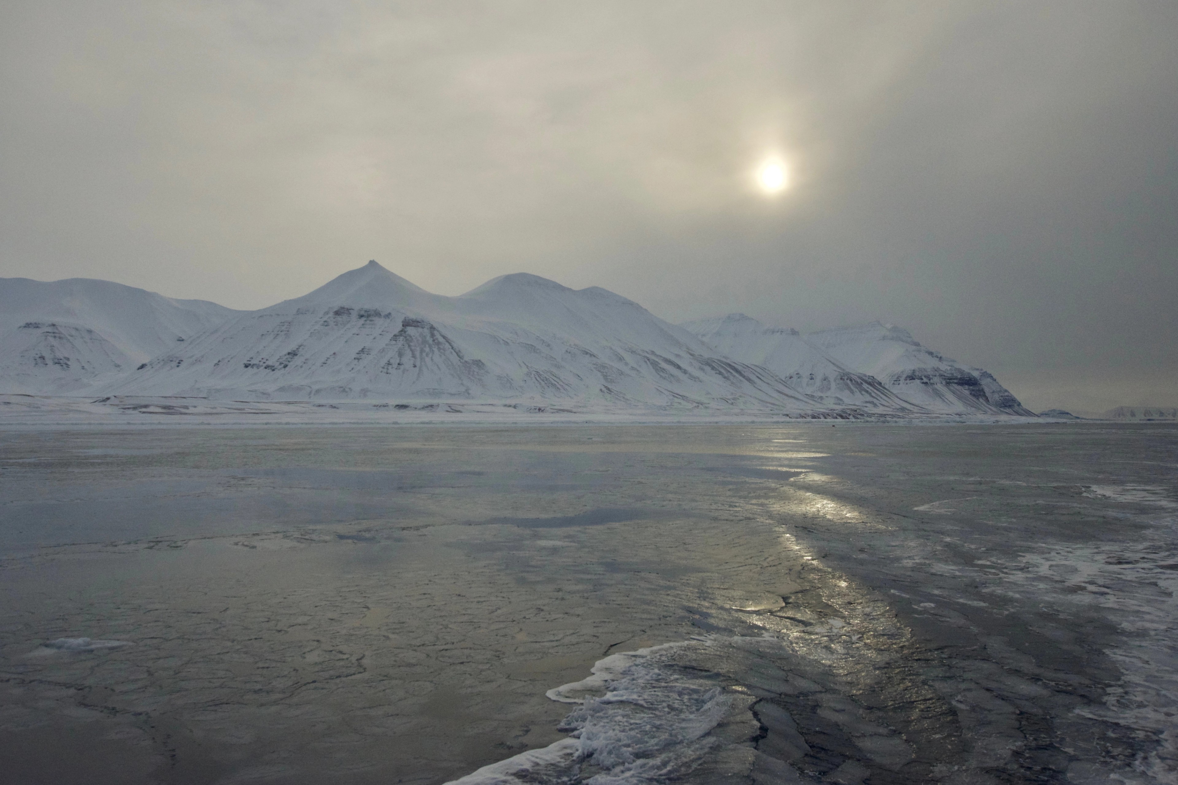 Low winter sun illuminates a frozen bay and snowy mountains.