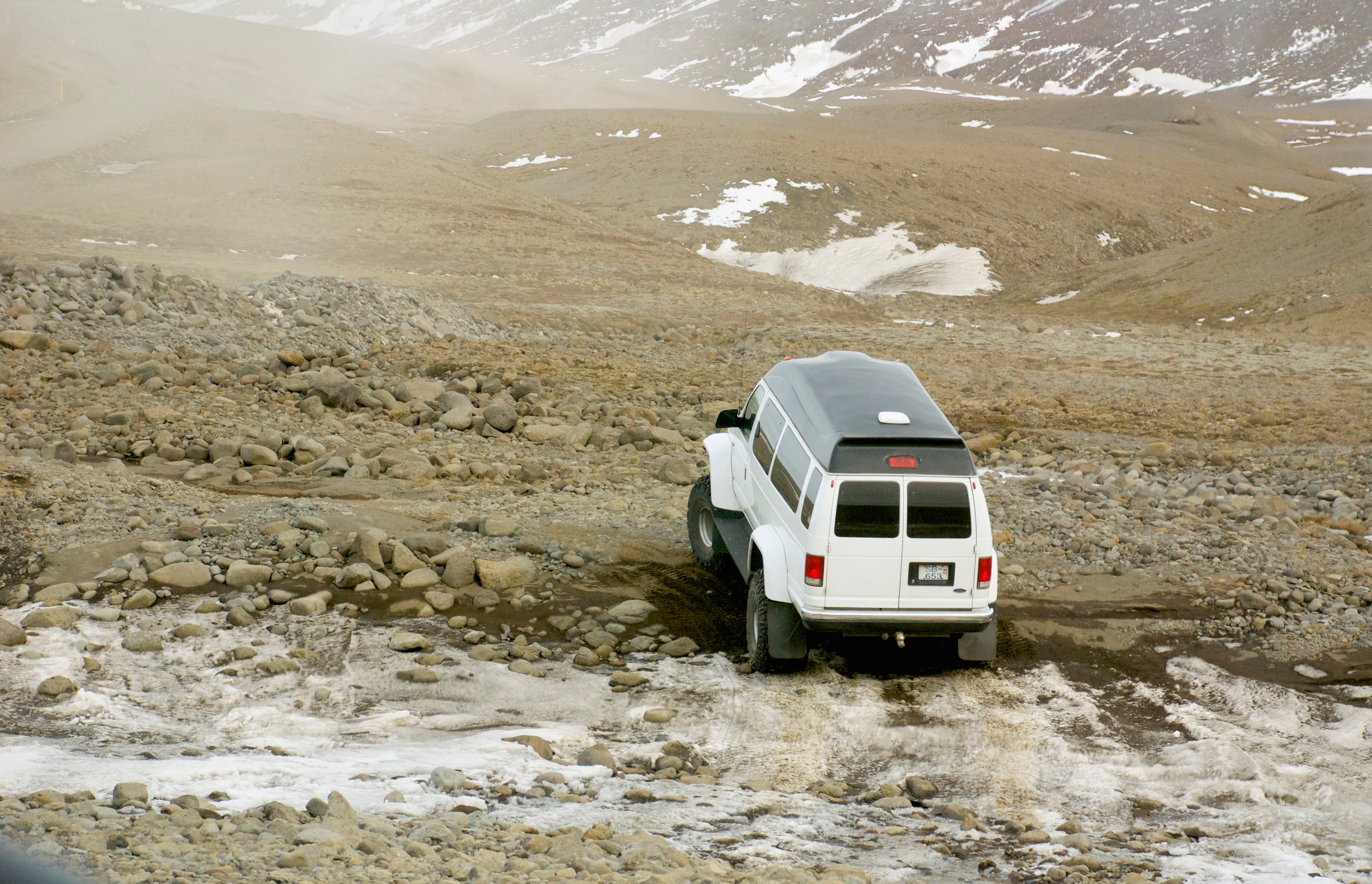 A white four-wheel-drive vehicle crosses a shallow stream on a rocky plain.