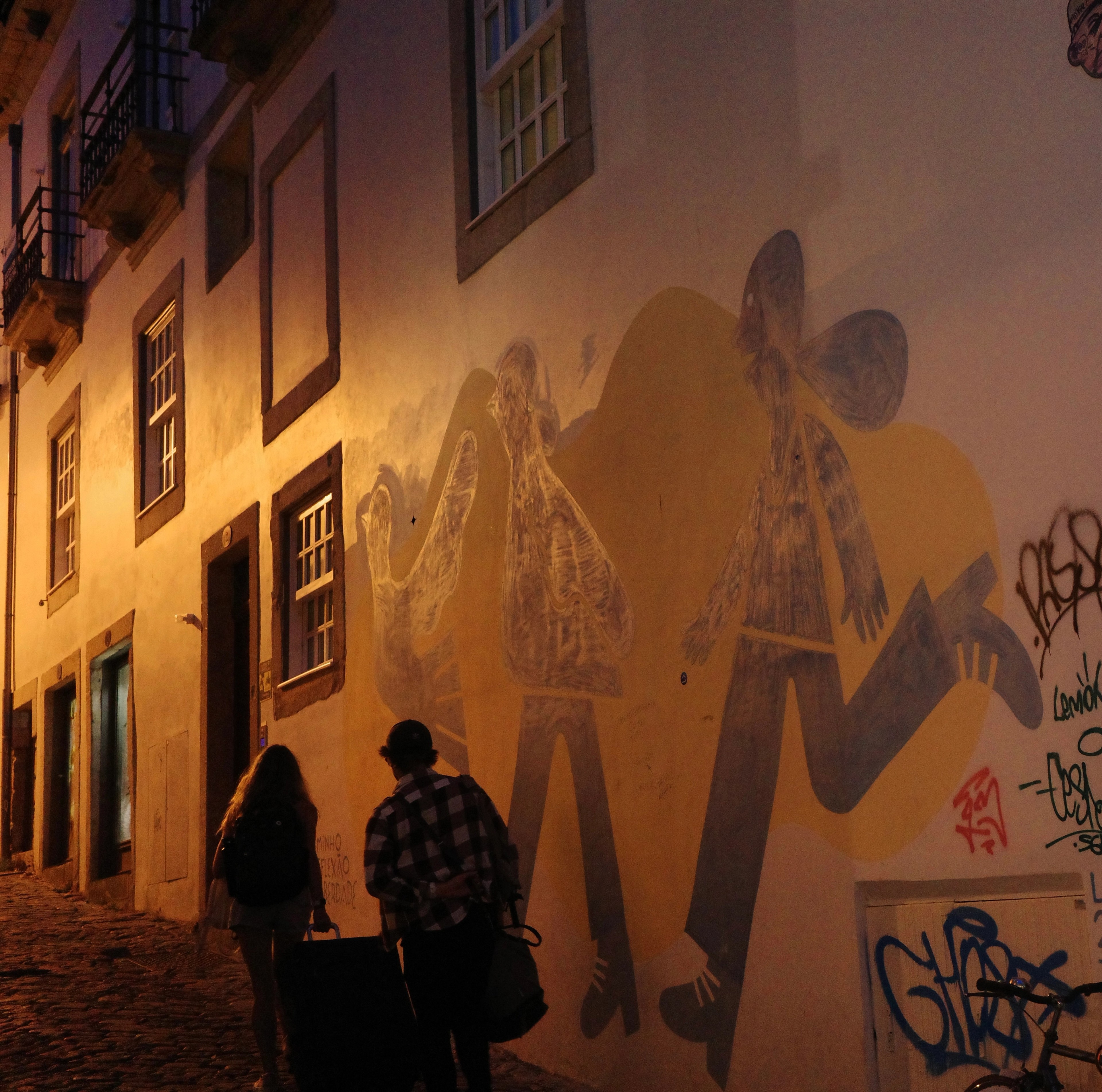 Two people look at a large illustrated mural on a warmly lit street wall.