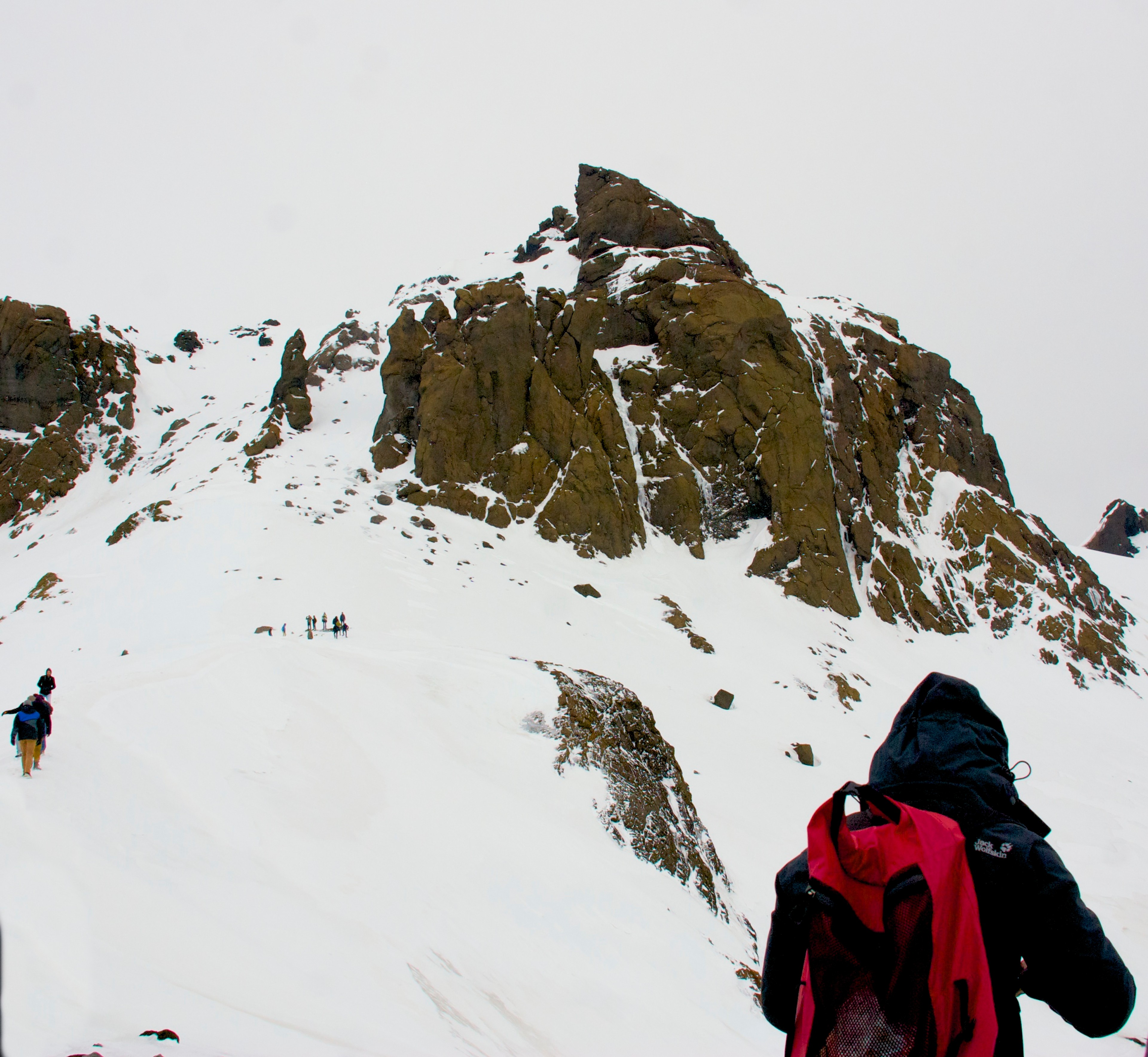 A person in a red jacket climbs across snow beside a dark rocky outcrop.