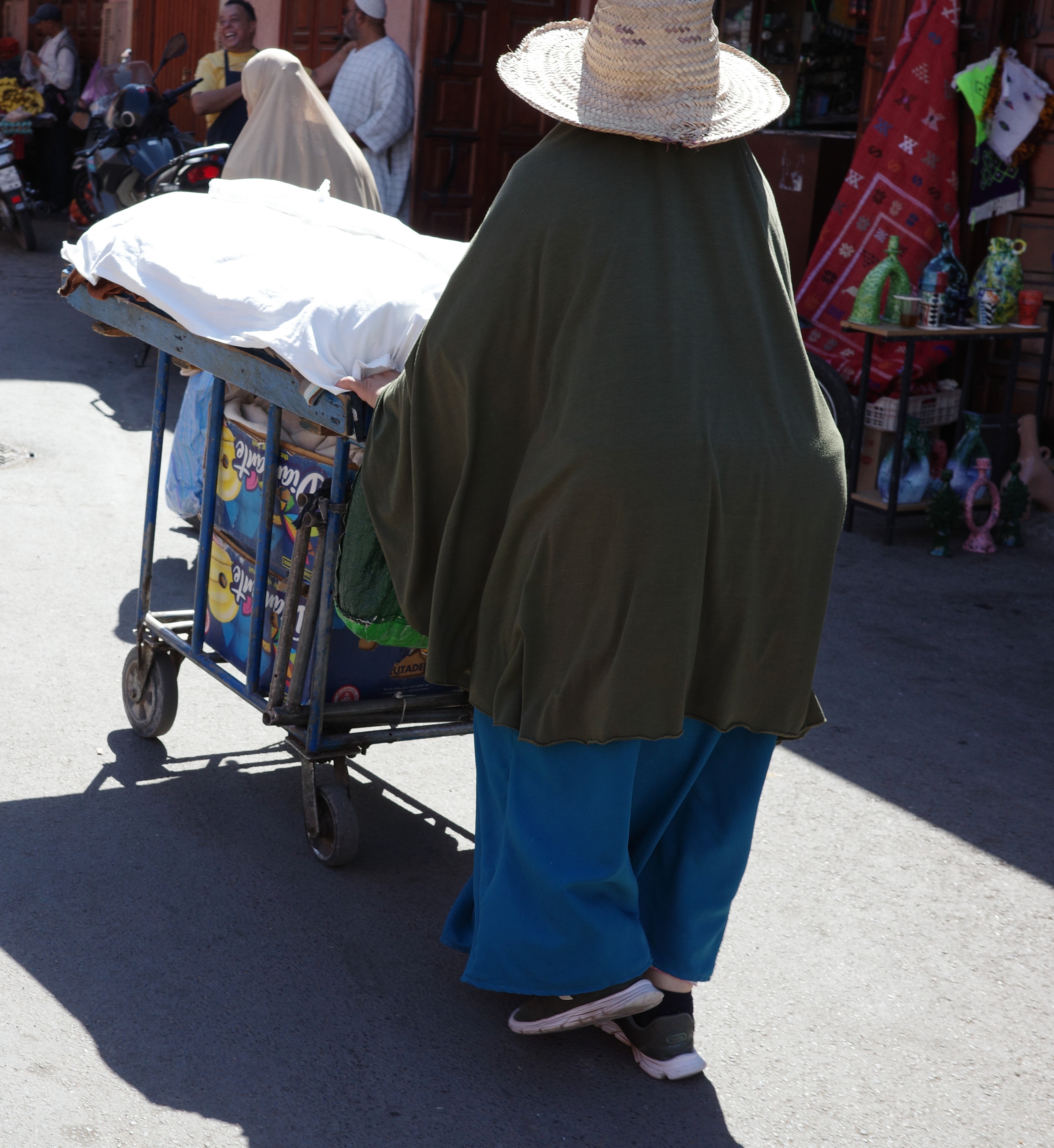 A person in a dark cloak pushes a cart through a busy market street.