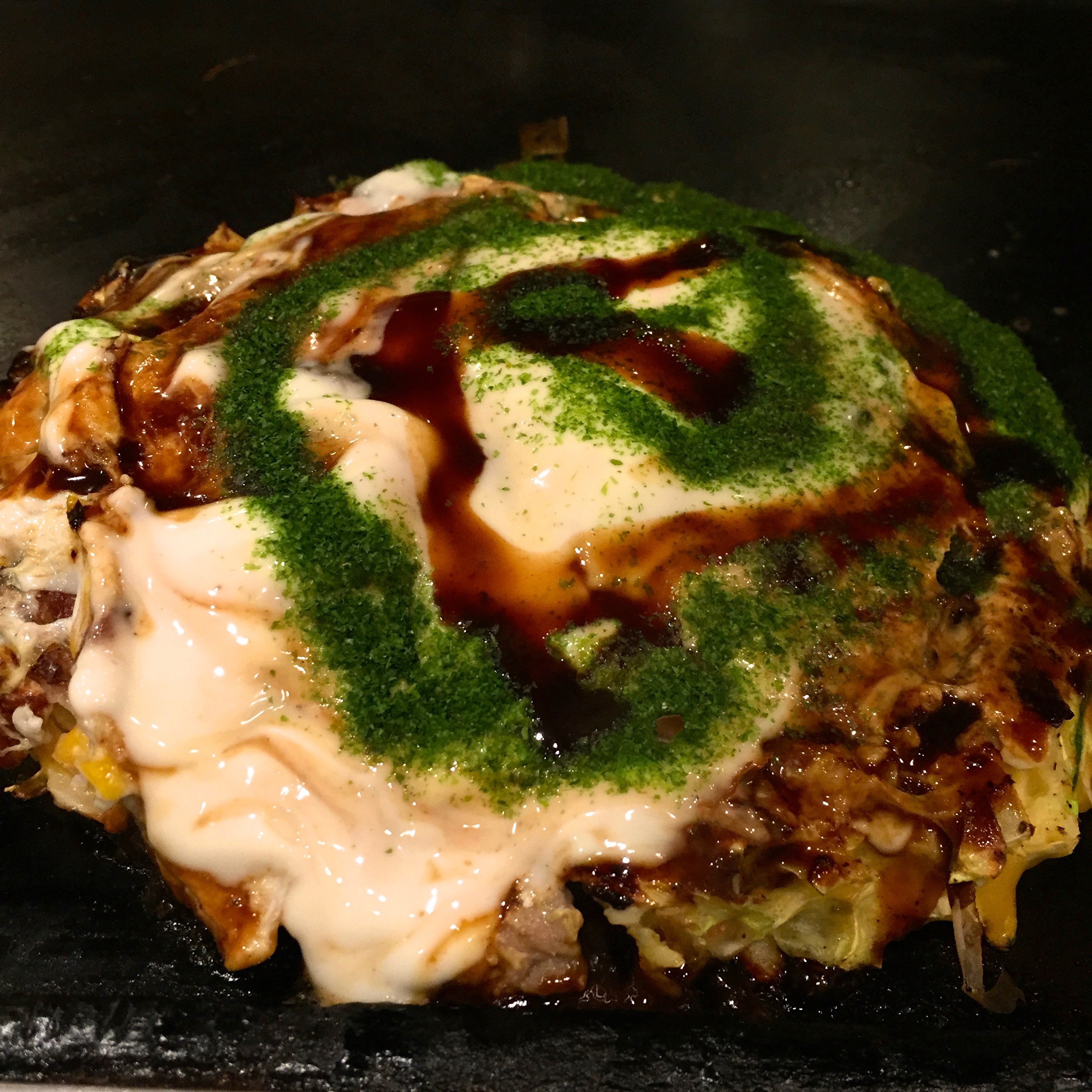 A savory pancake is topped with sauce, mayonnaise, and green garnish.