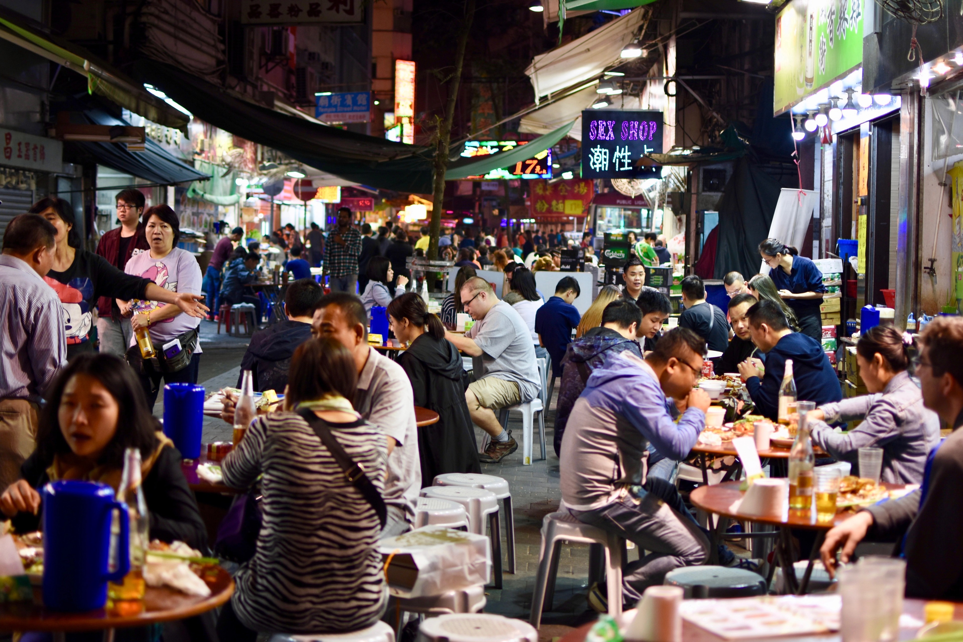 Diners sit at crowded sidewalk tables in a busy night market street.