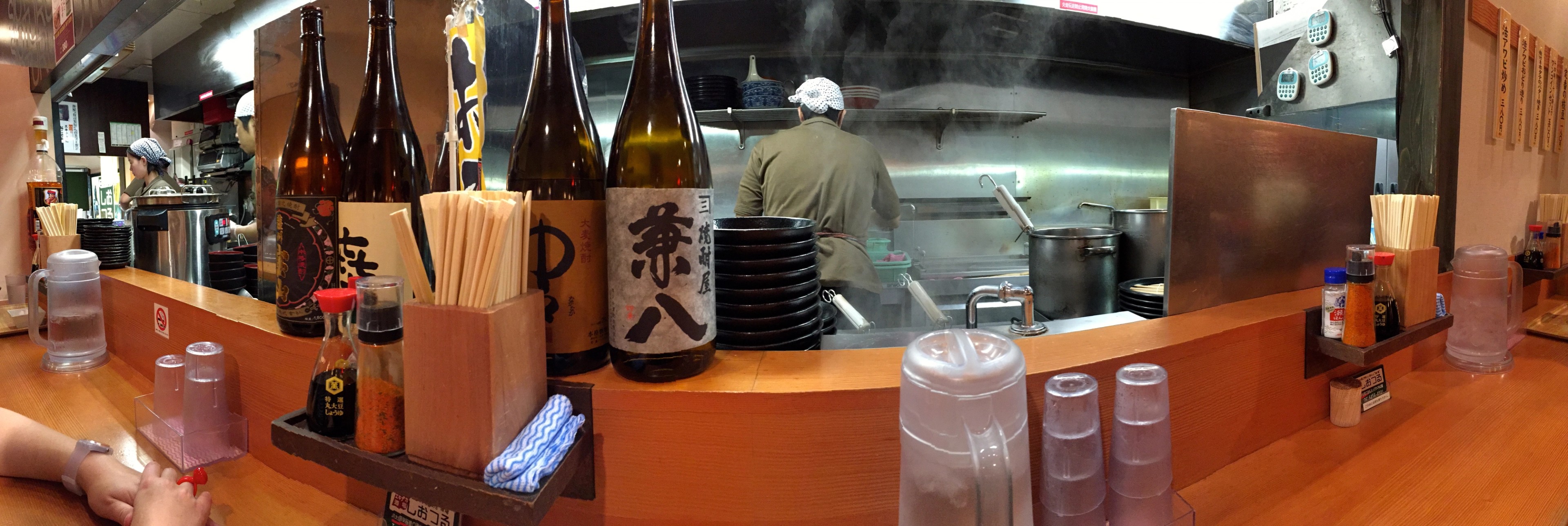 A wooden restaurant counter is lined with bottles, cups, and cooking equipment.