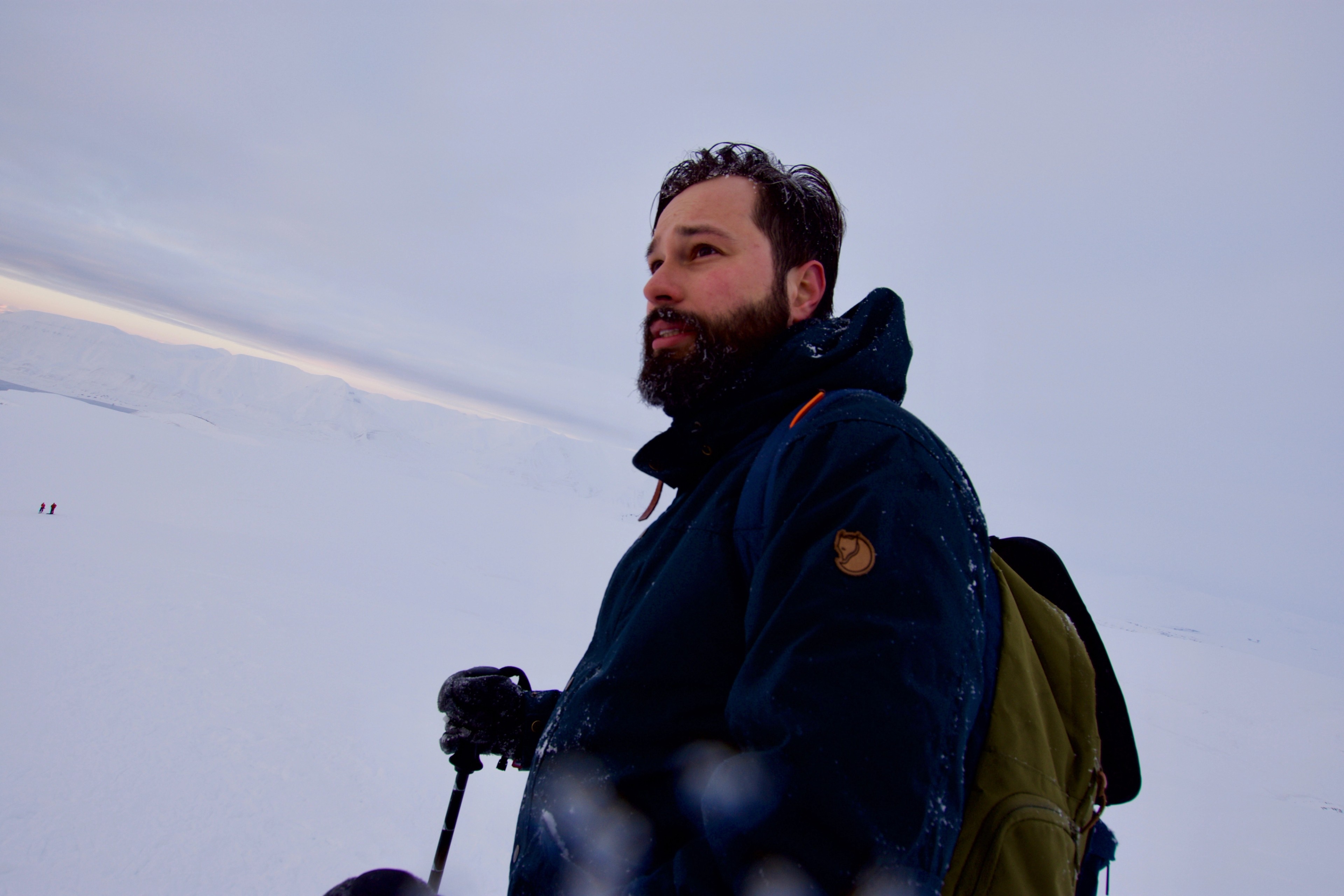 A bearded hiker stands in a dark jacket against the snow.