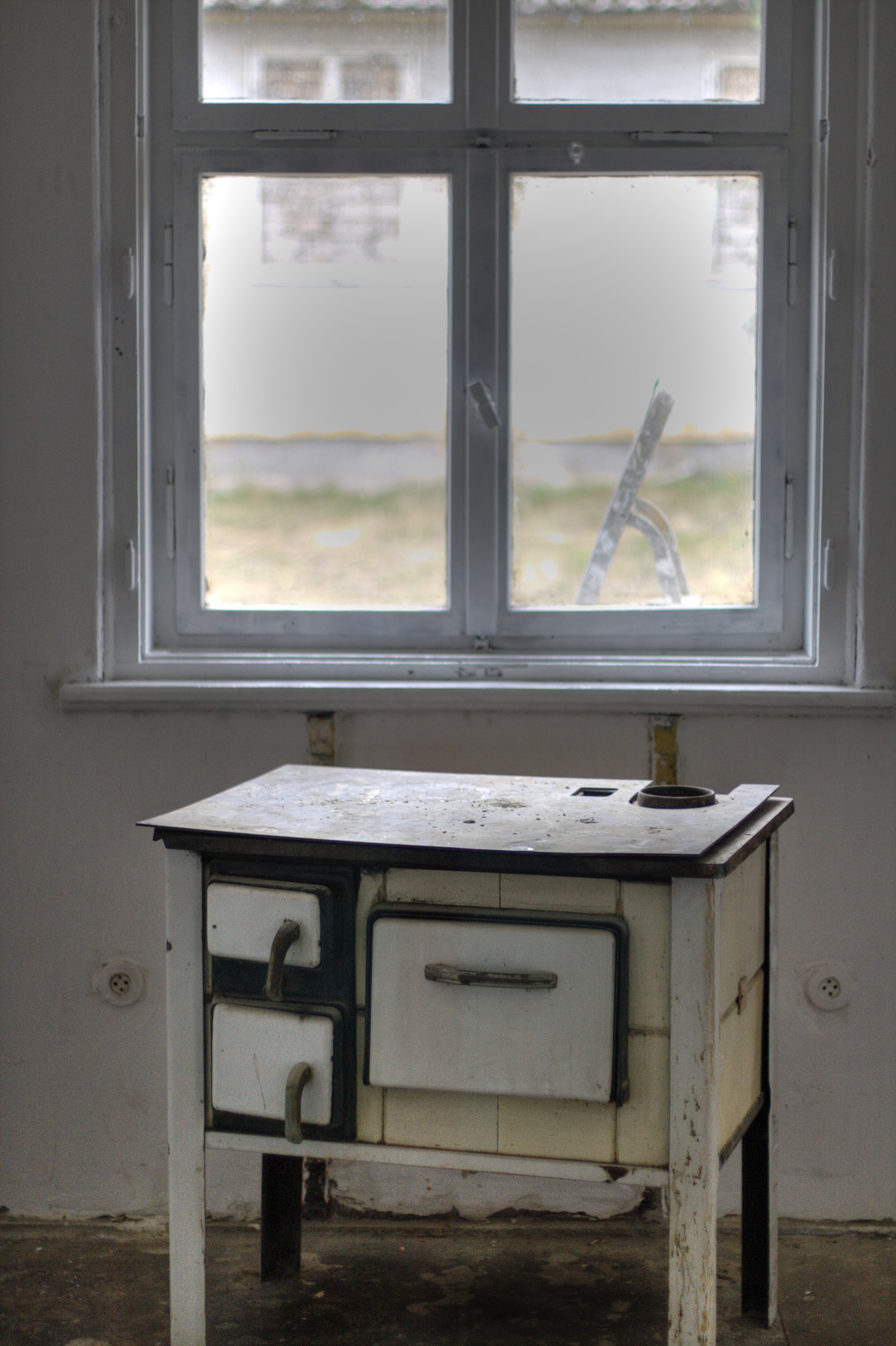 A small old stove stands below a window in a bare room.