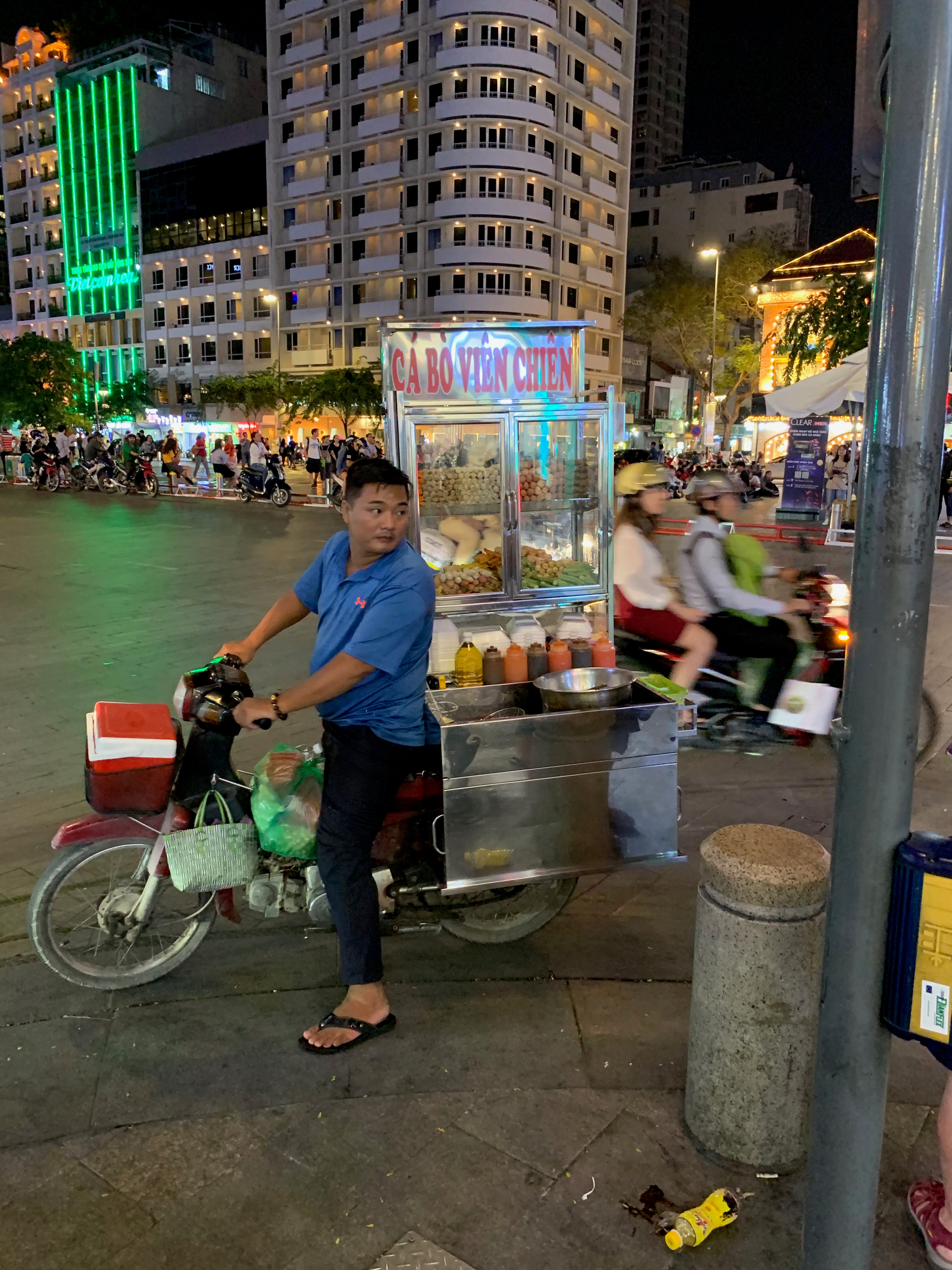 A man stands beside a bicycle cart in a brightly lit night street.