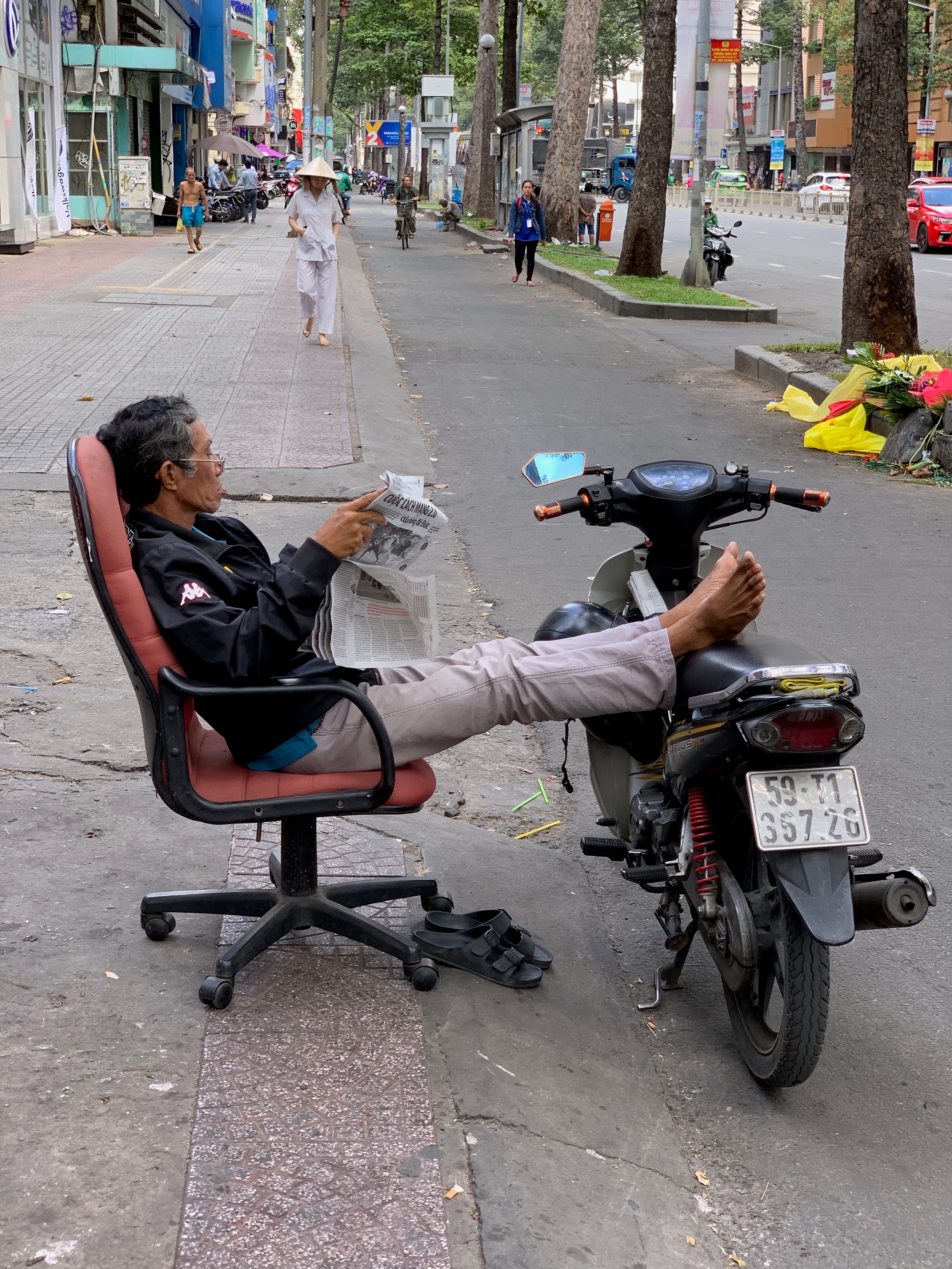 A man reclines on a chair attached to a parked motorbike at the curb.