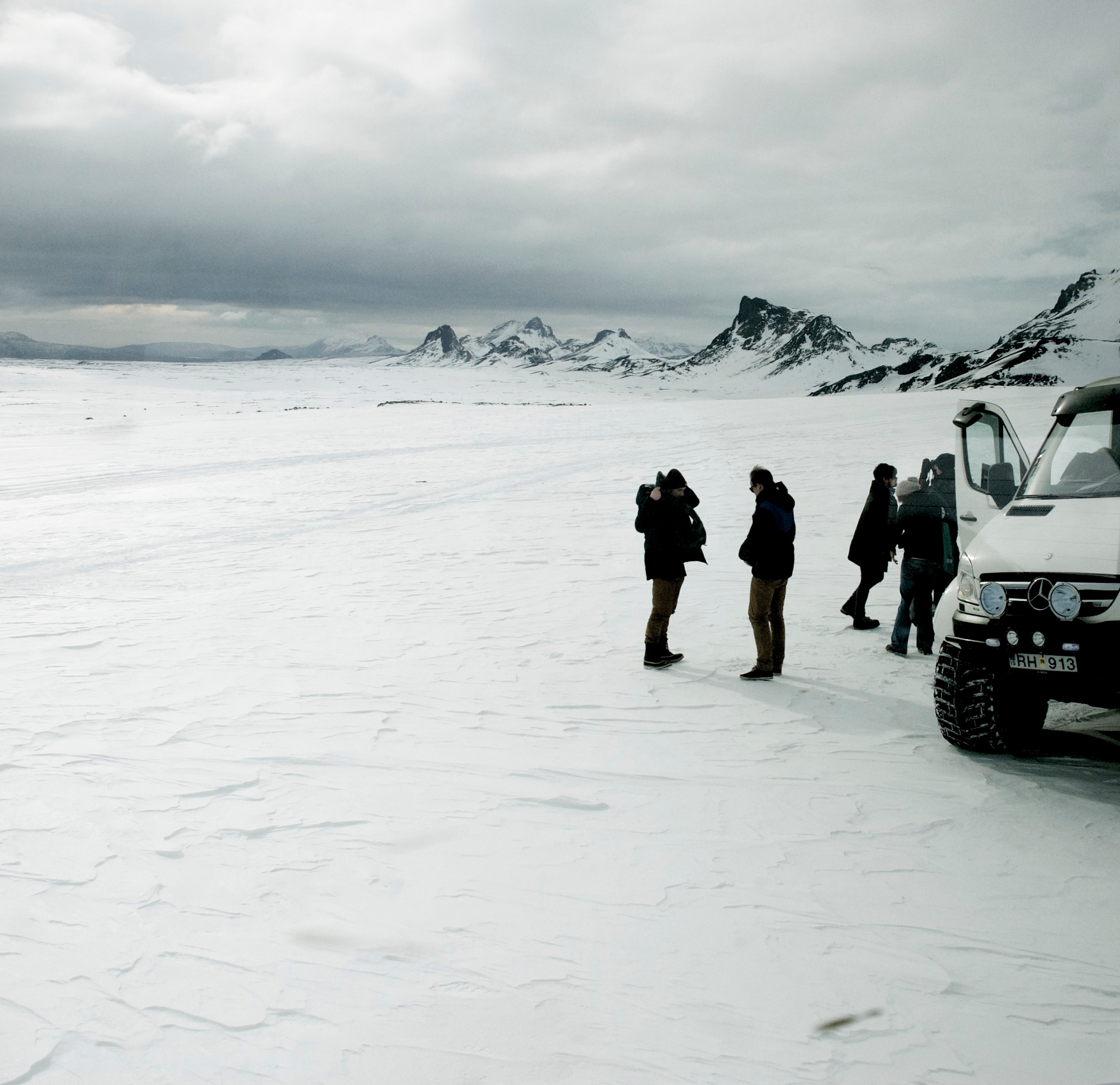 Hikers and vehicles stand on a snowfield beneath steep mountain peaks.