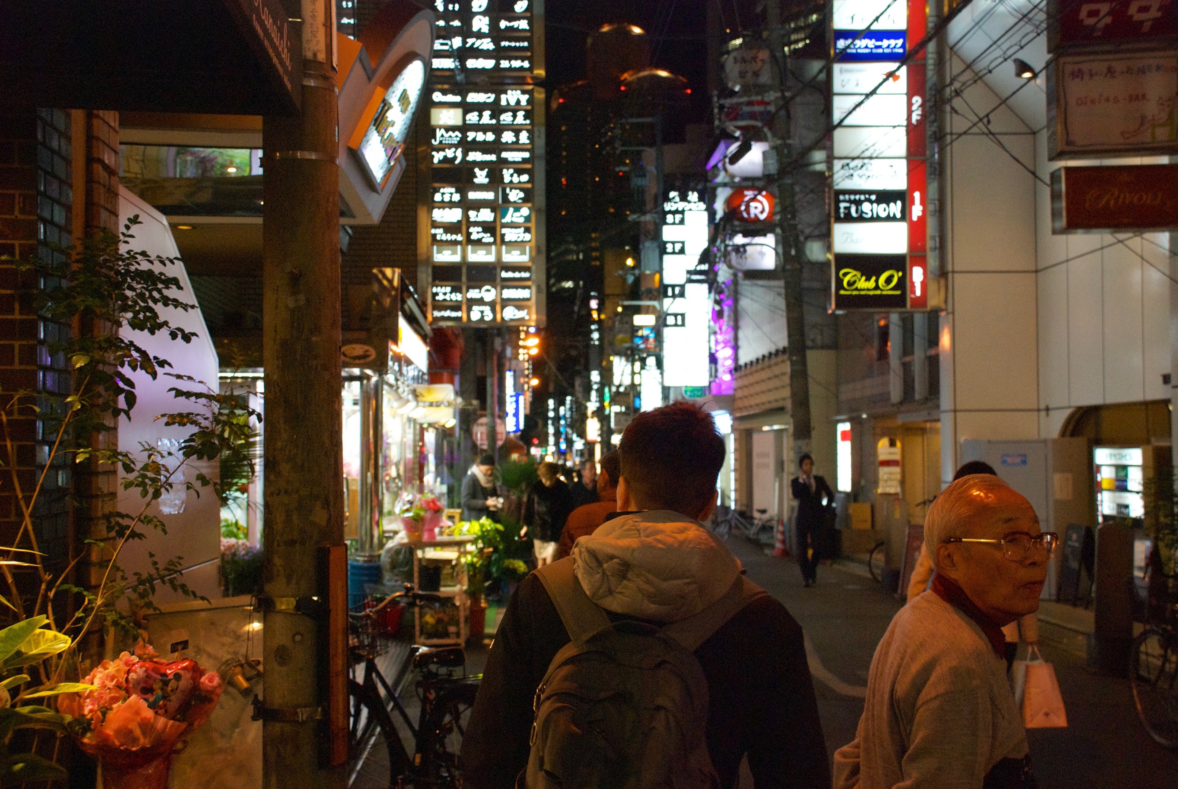 Pedestrians walk through a brightly lit city street at night.