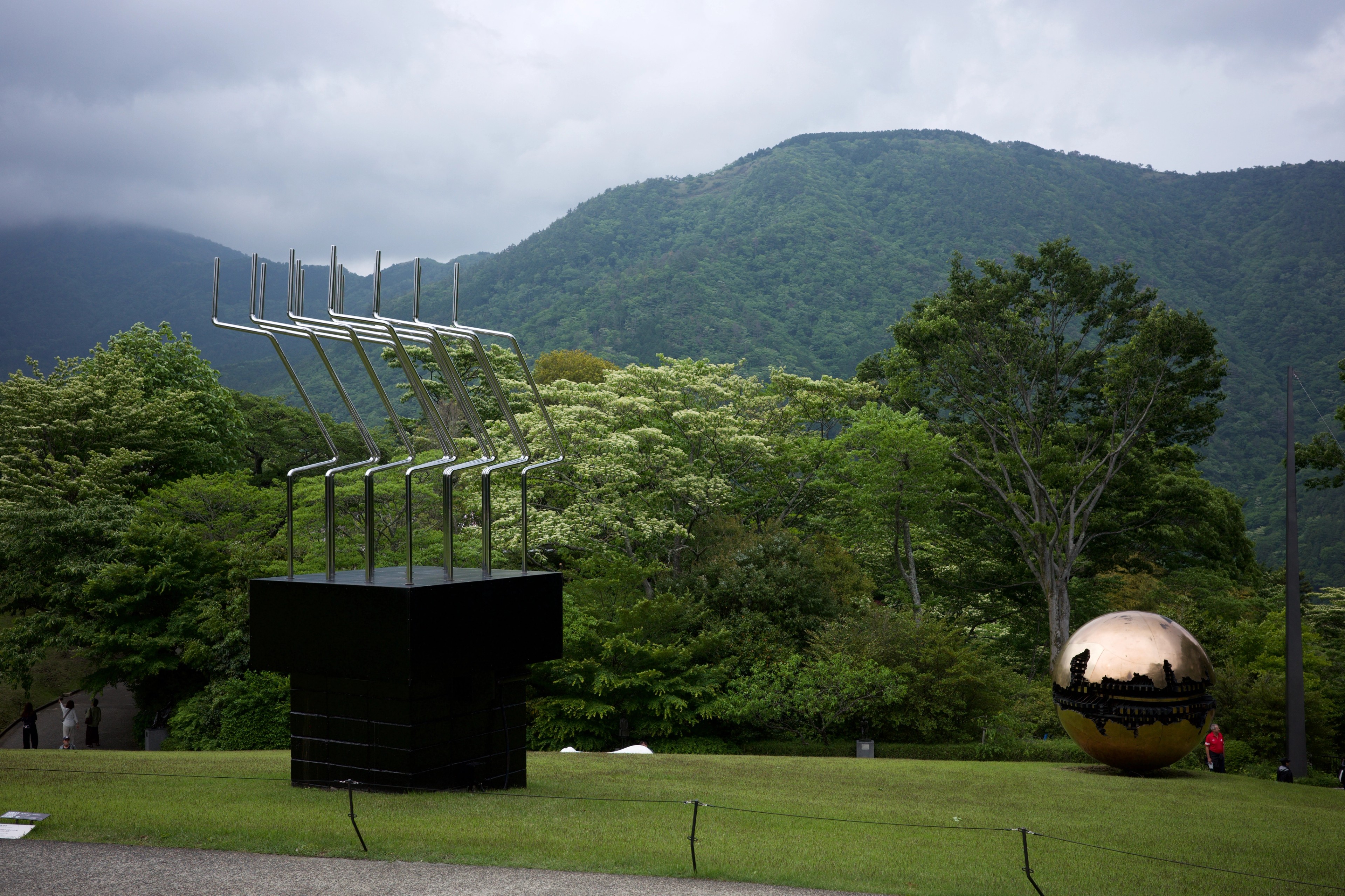 A green park beneath a cloudy mountain ridge with a dark sculpture.