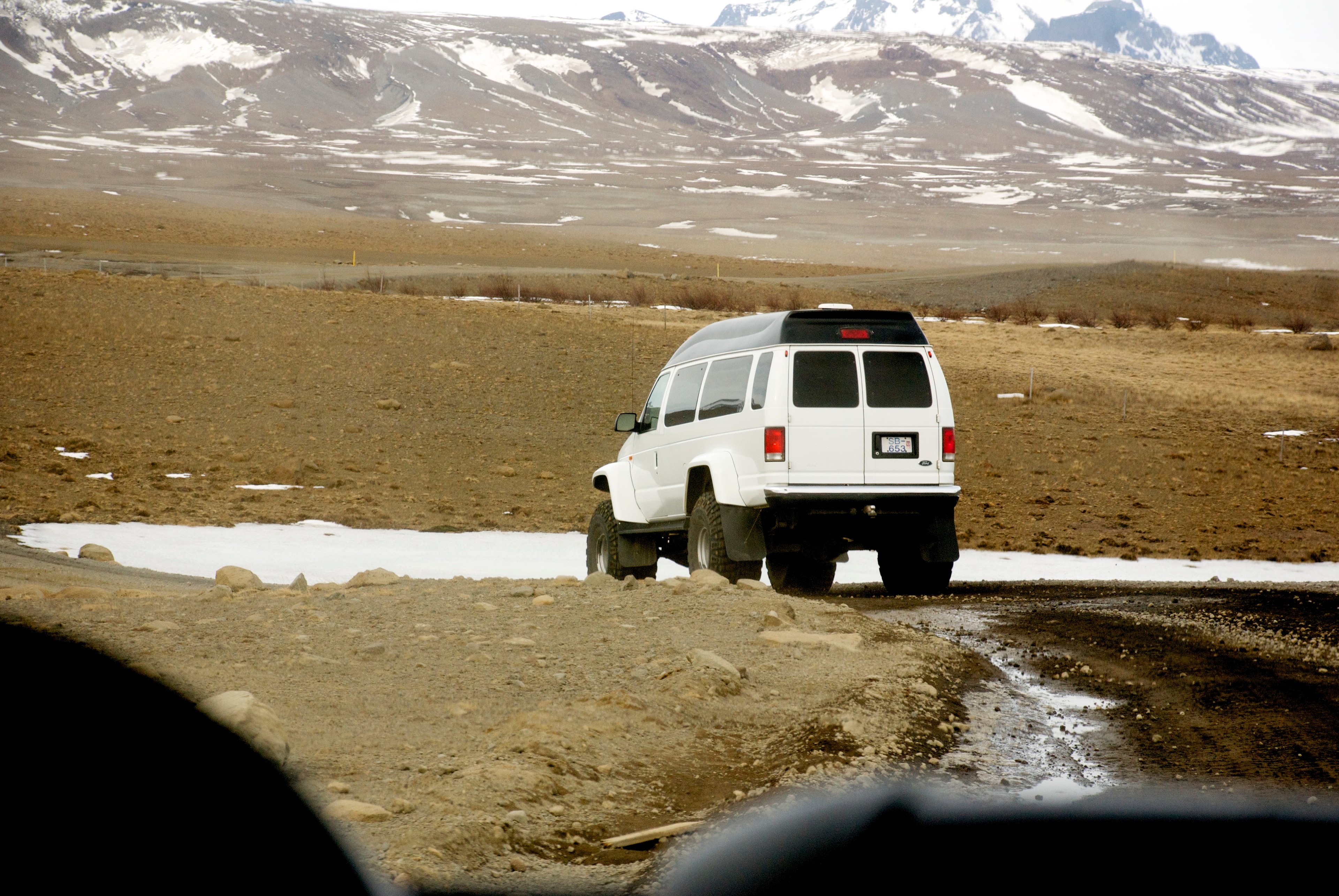 A white four-wheel-drive vehicle travels across a broad brown plain beneath snowy mountains.