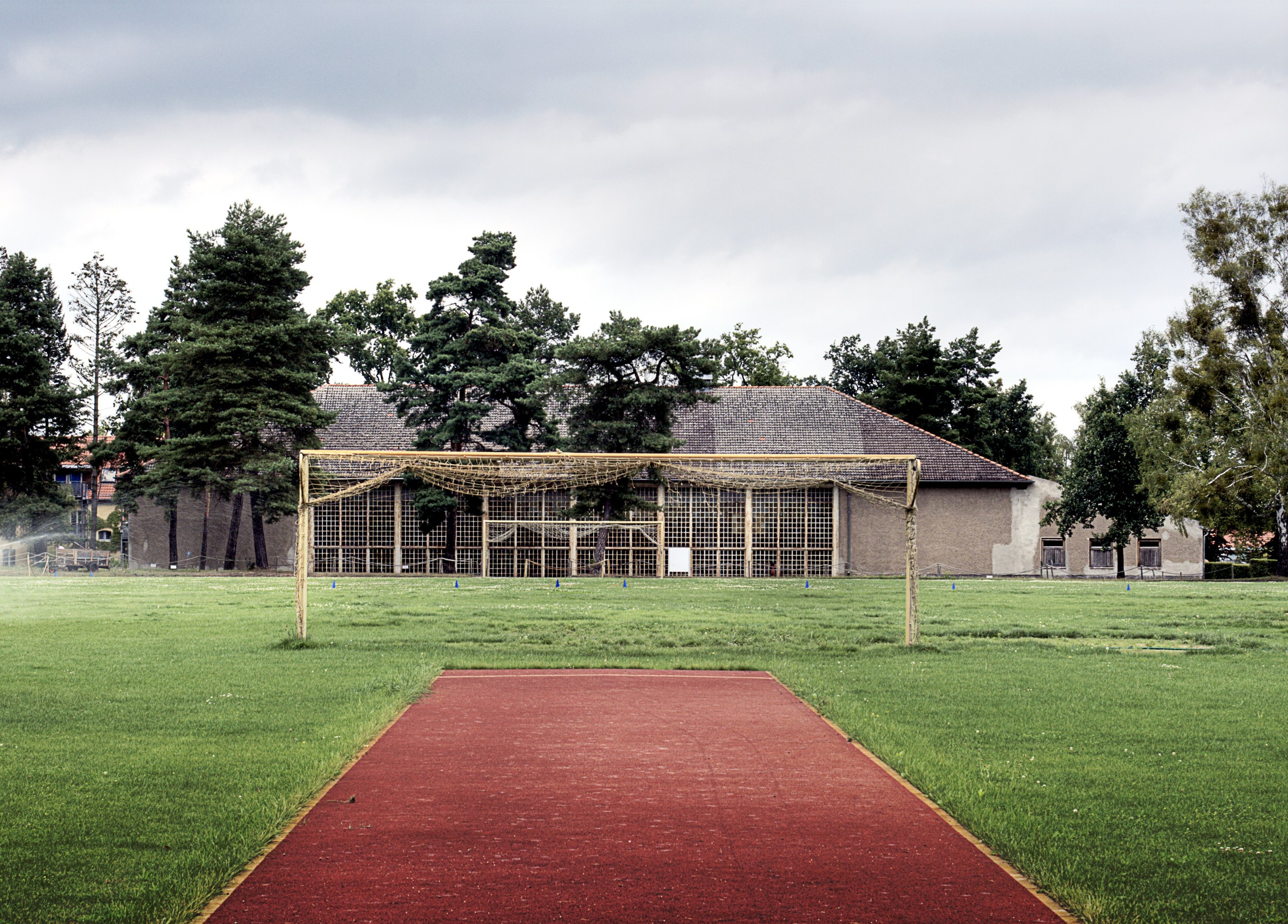 A red running track crosses a grassy field toward a low, weathered building.