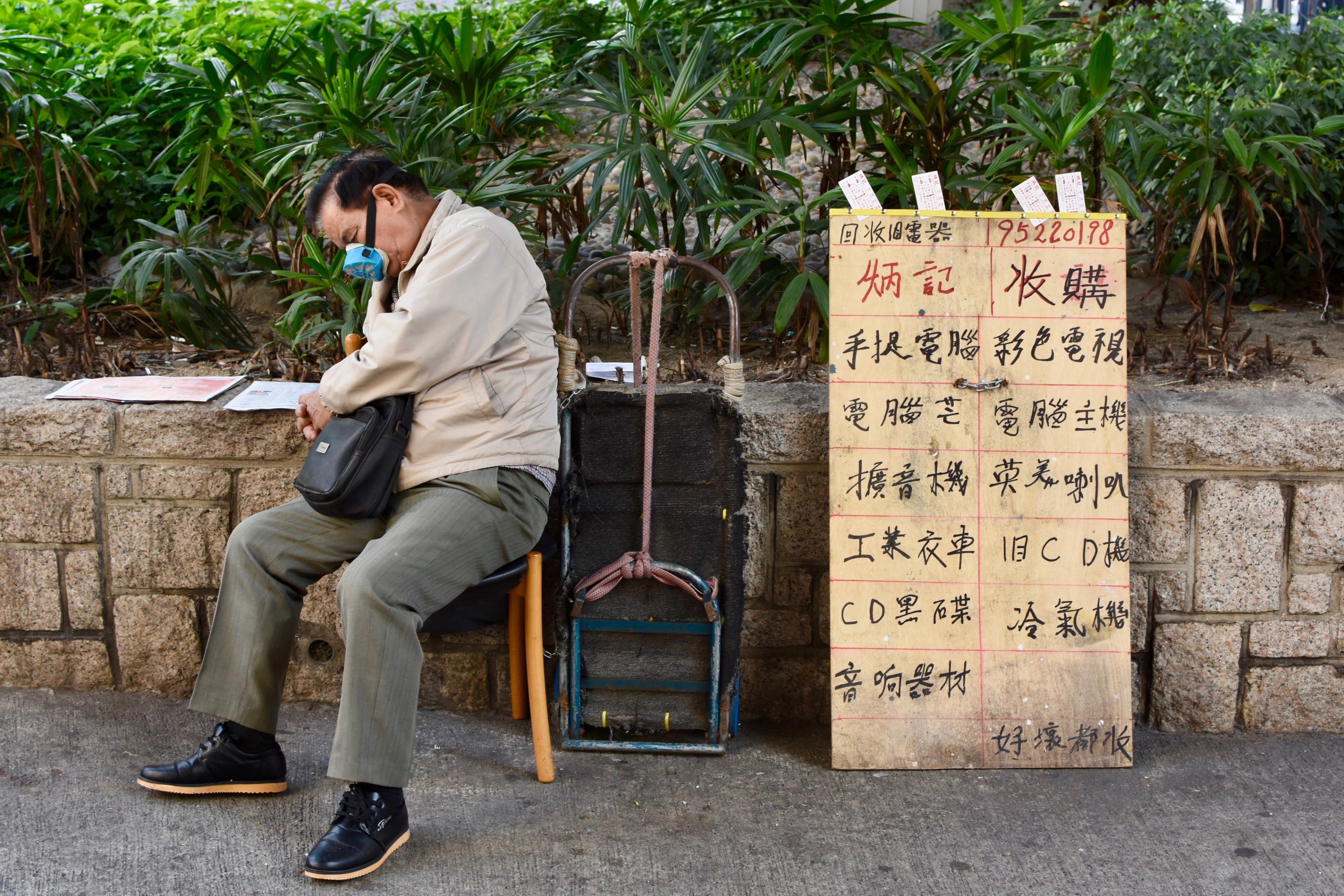 A seated man rests beside a handwritten sign in a public area.