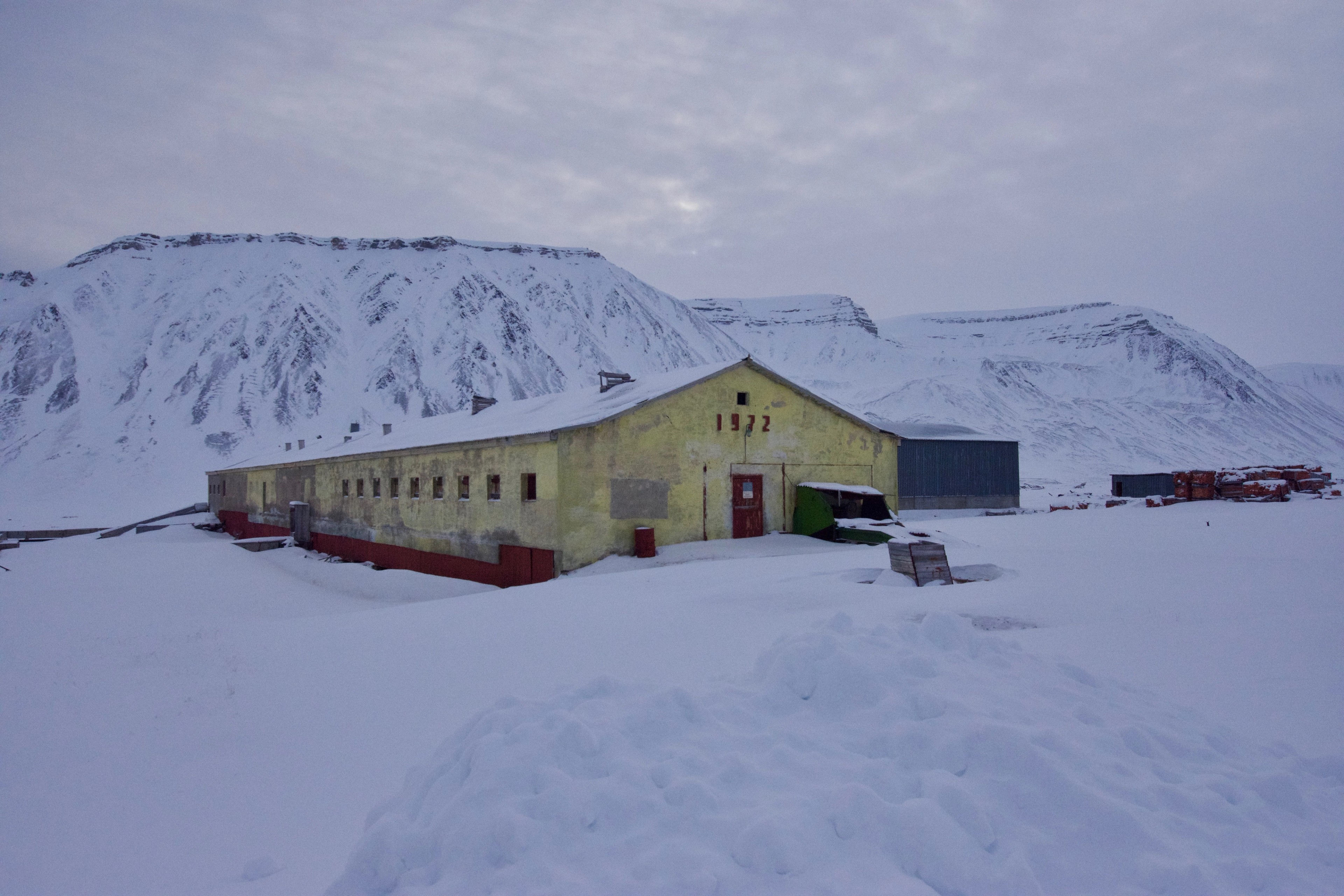 A yellow building stands in a snowy settlement below a mountain.