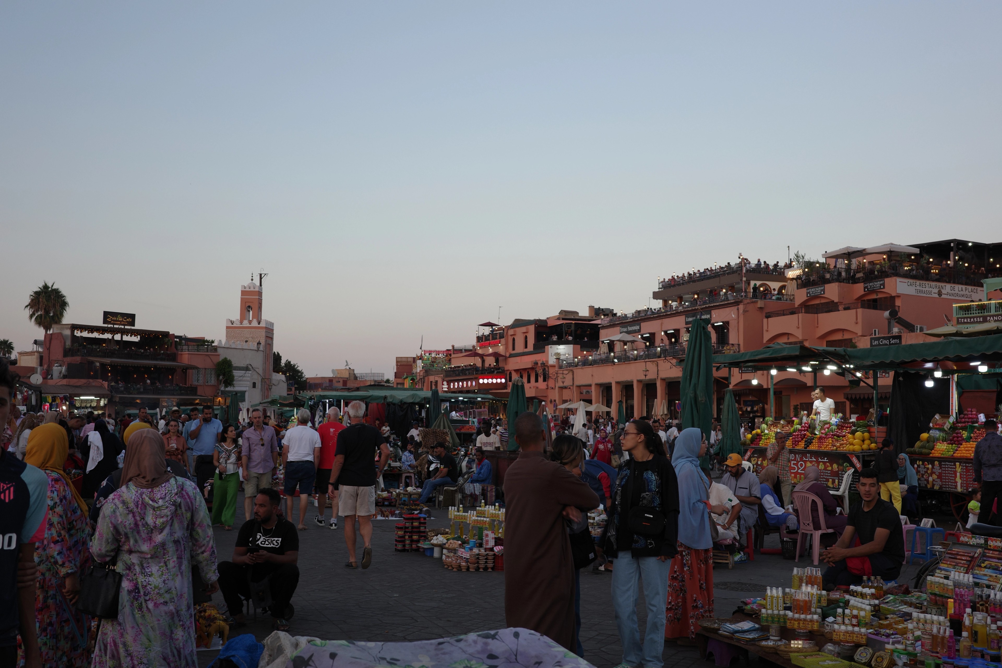A crowded market square is filled with people and stalls beneath the evening sky.