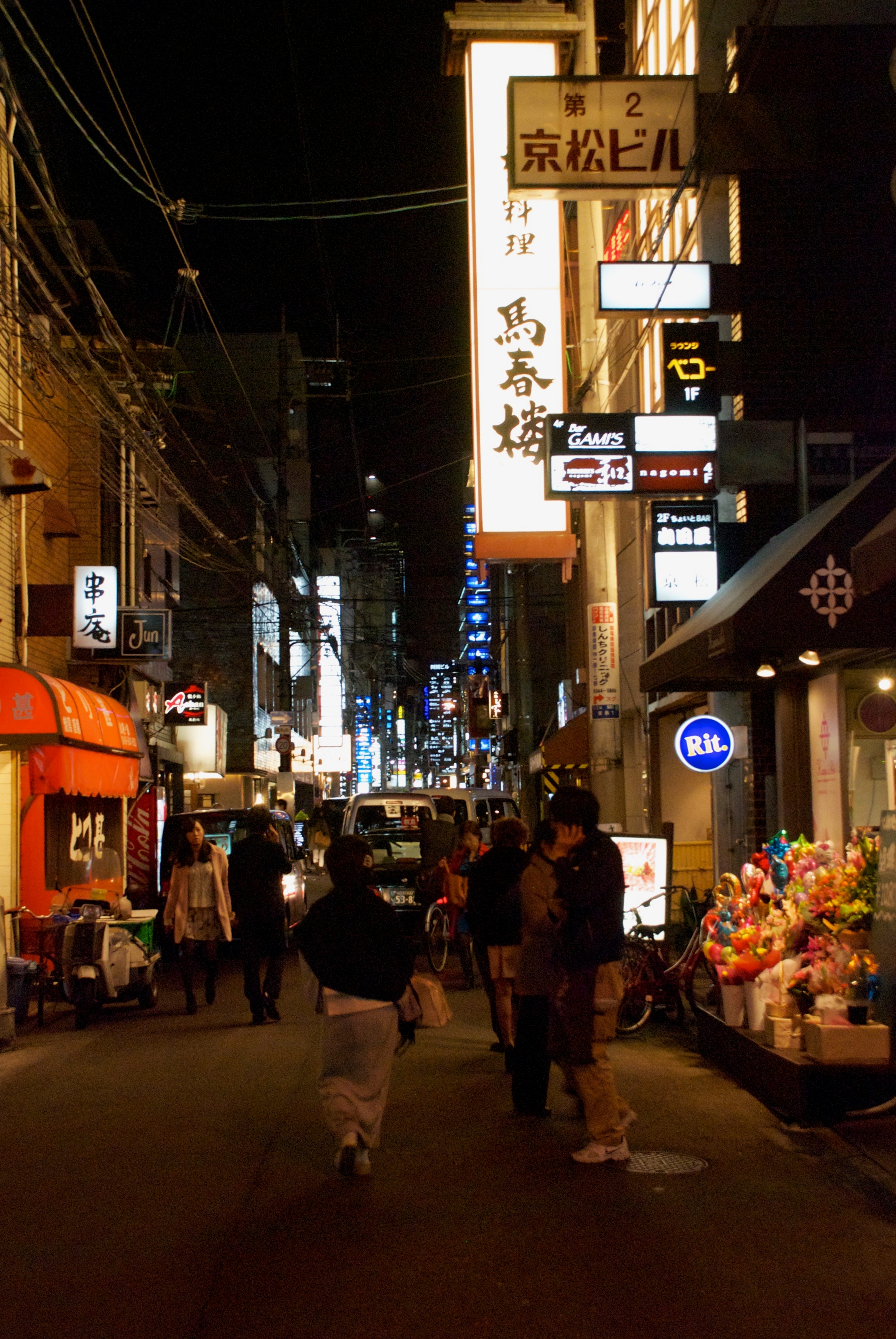 A narrow commercial street is crowded with illuminated signs after dark.