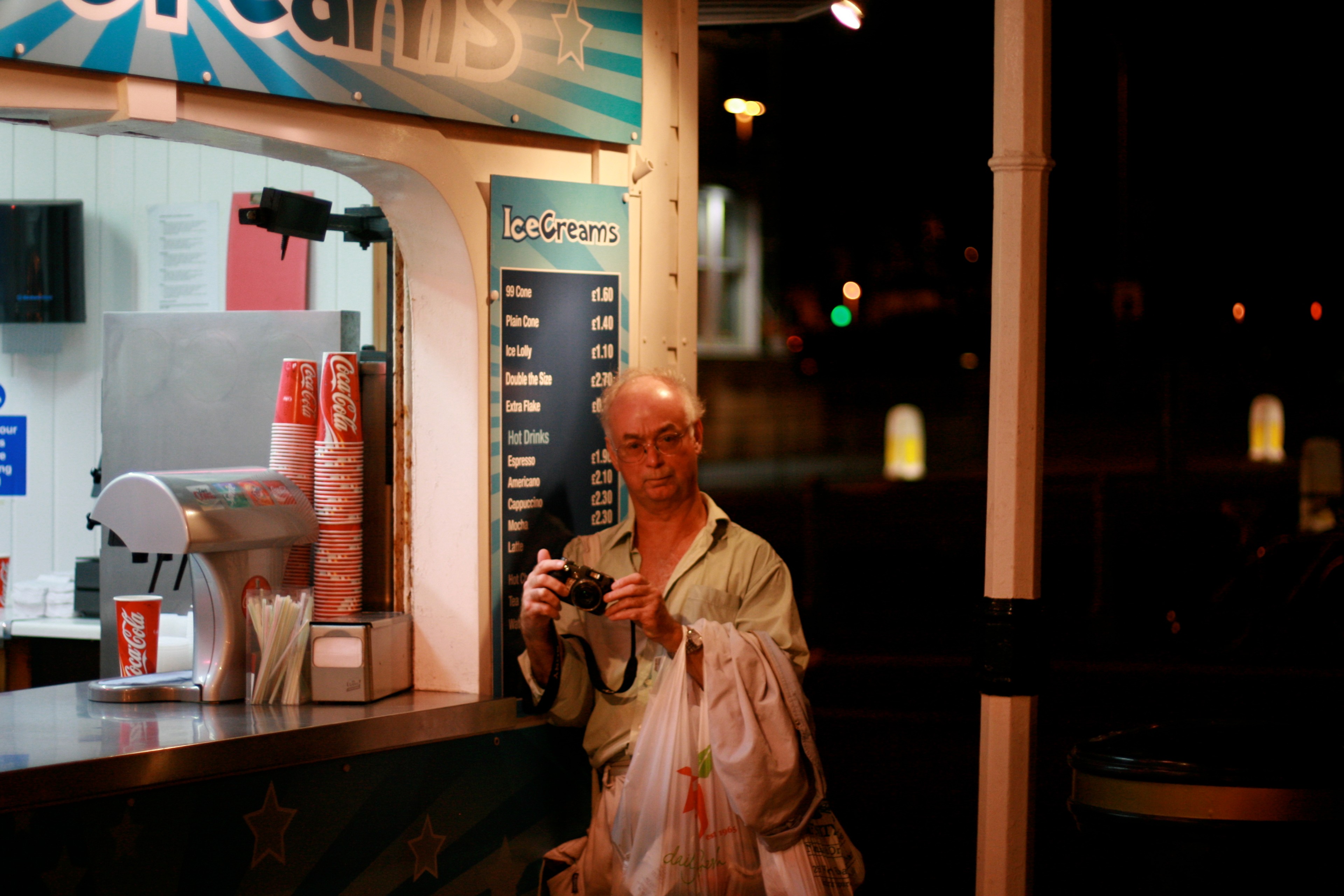 An elderly man stands beside a brightly lit takeaway counter at night.
