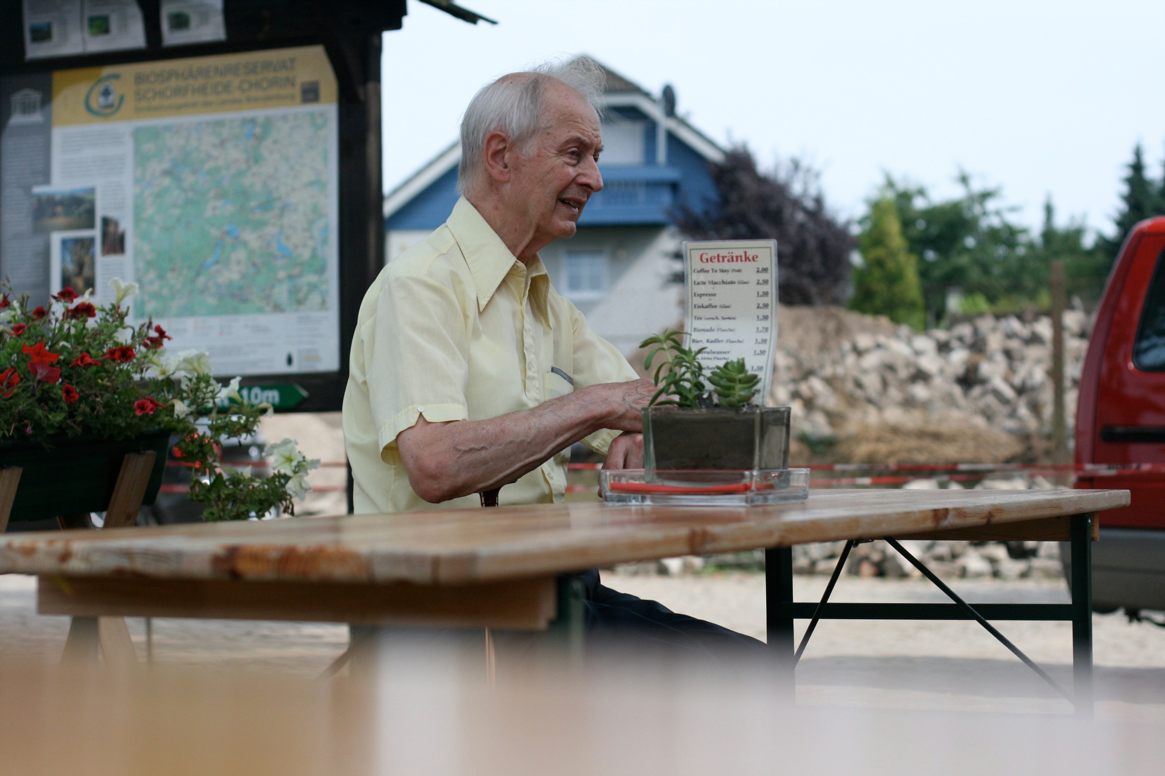 An older man stands behind an outdoor table and holds up a paper map or document.