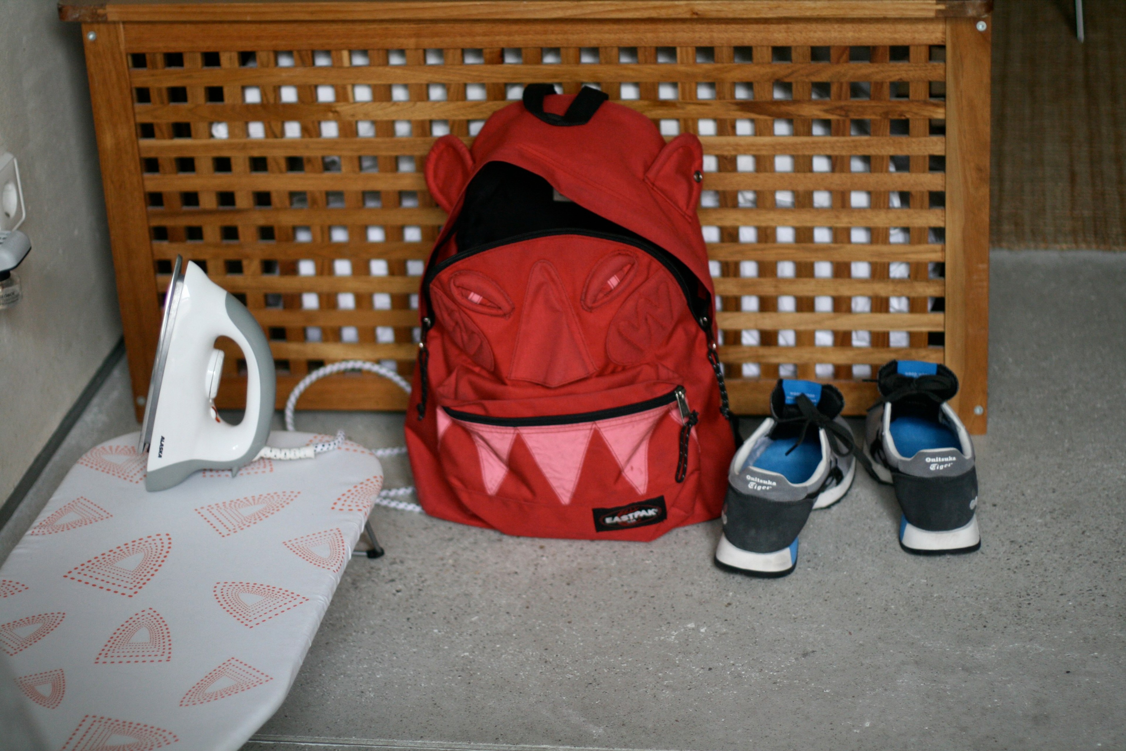 A red backpack, shoes, and a small iron are arranged on a tiled floor beneath a wooden lattice.