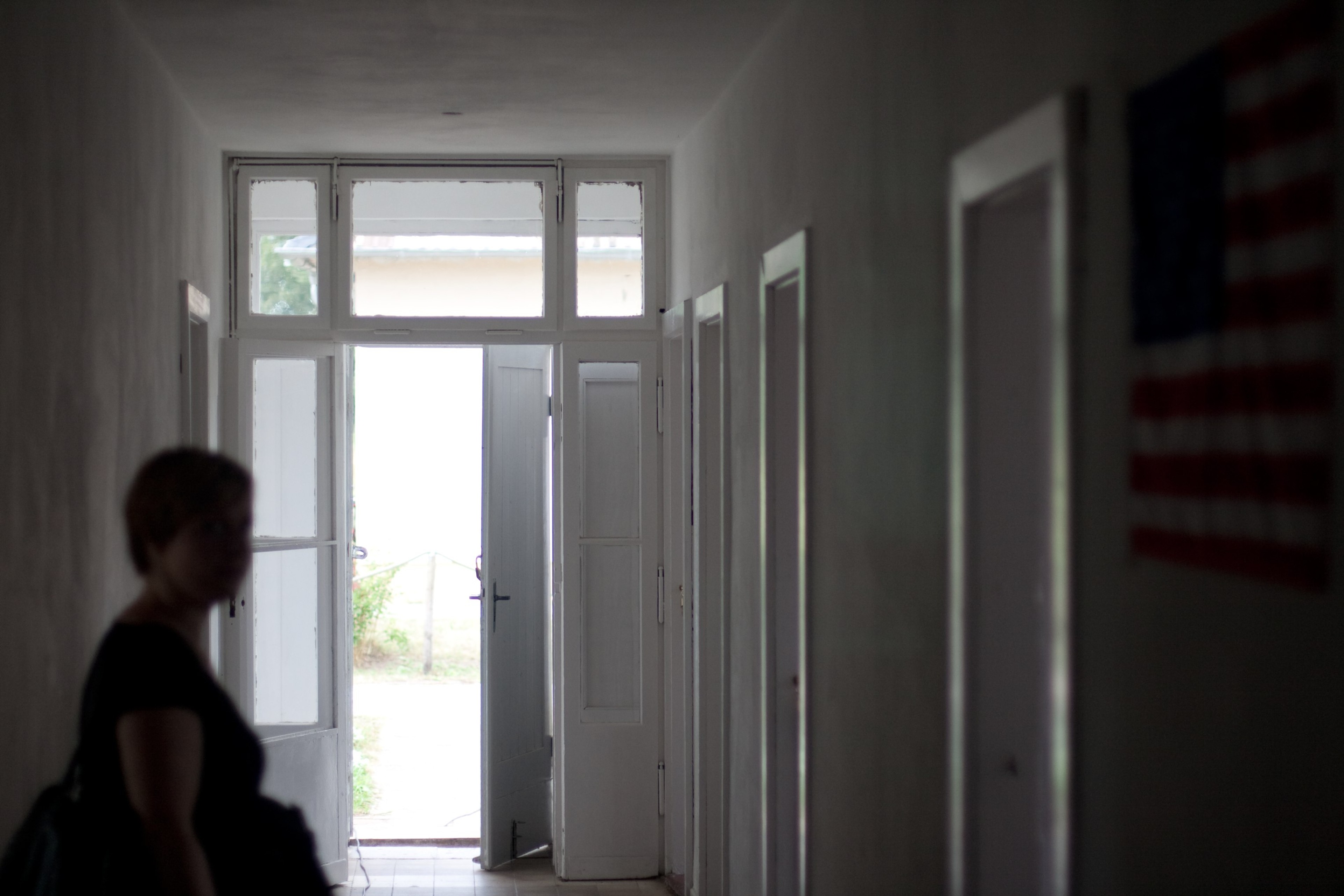 A person stands in silhouette beside a bright doorway in a dim hallway.