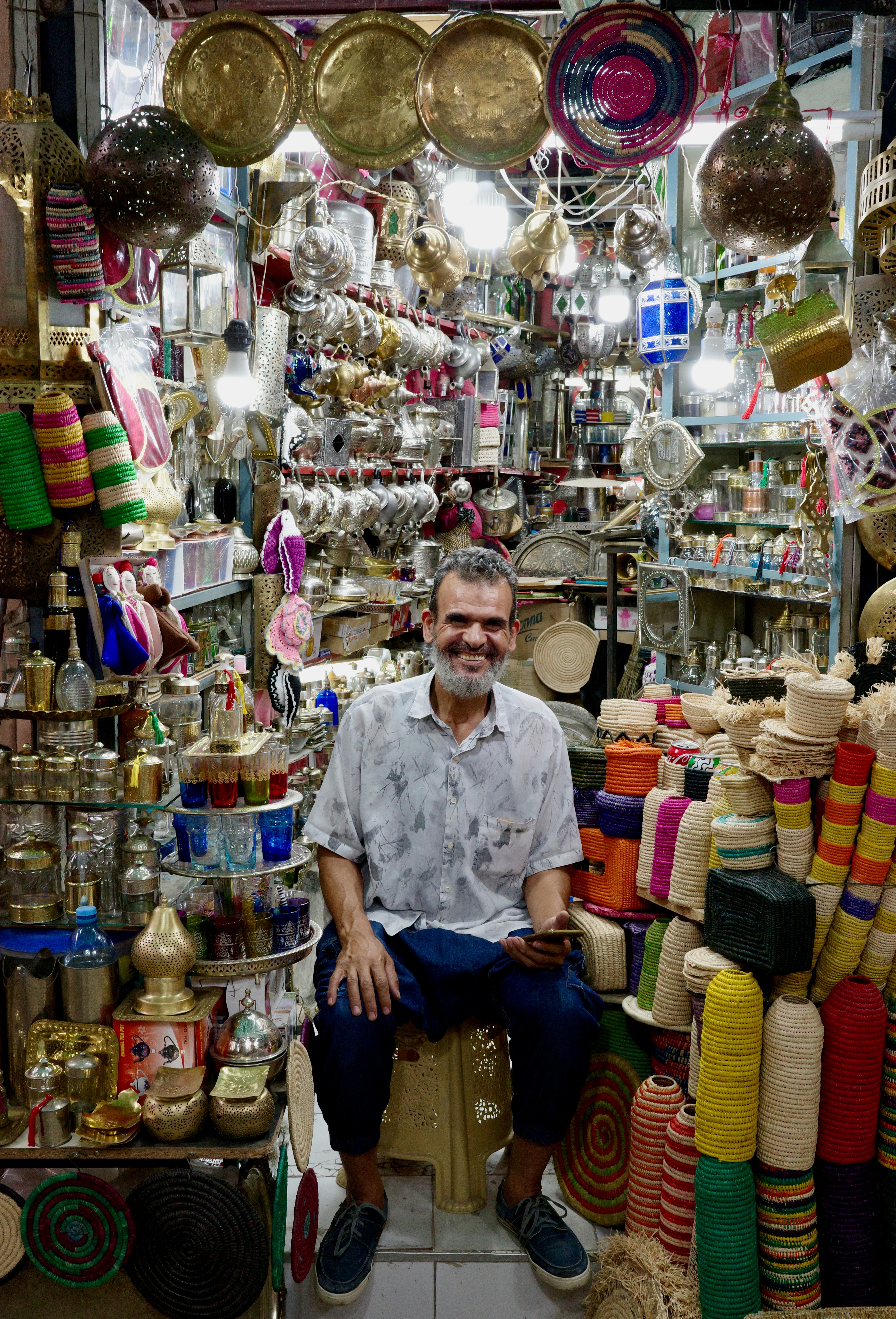 A shopkeeper sits among hanging metal lamps and densely packed market goods.