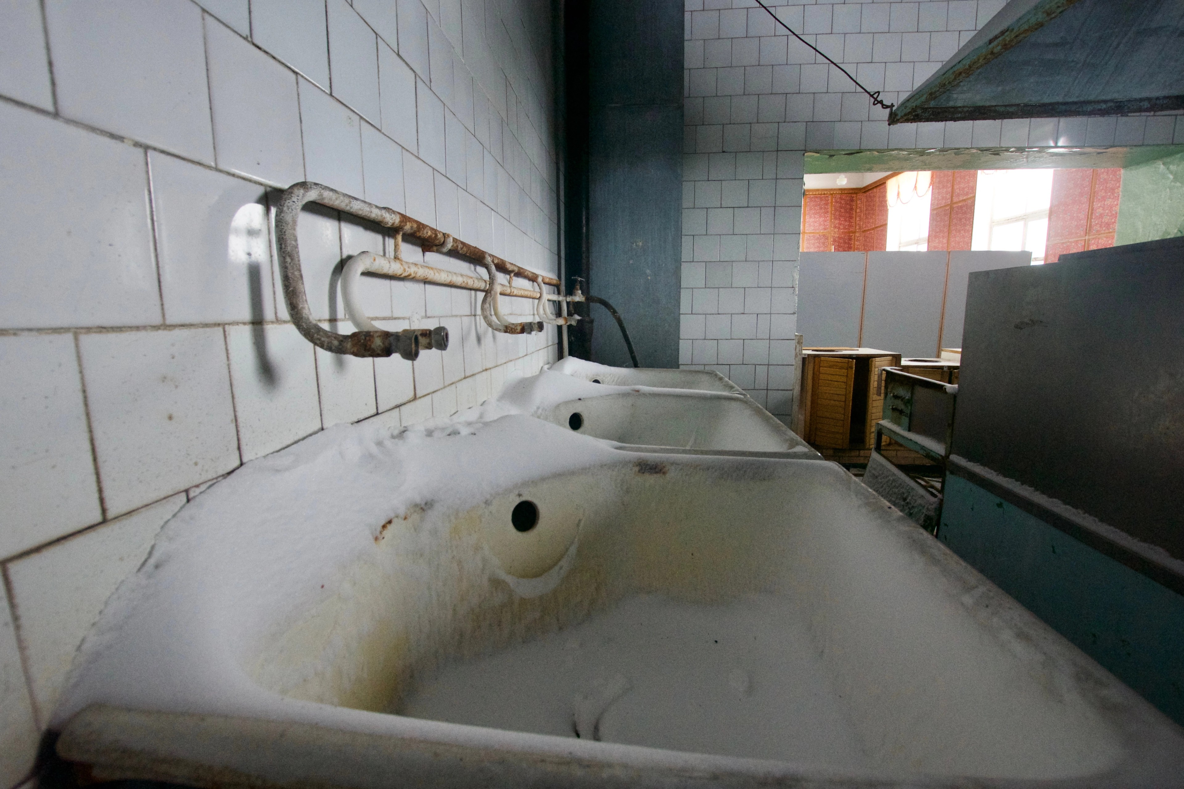 A bathtub and pipes in a deteriorated tiled bathroom.