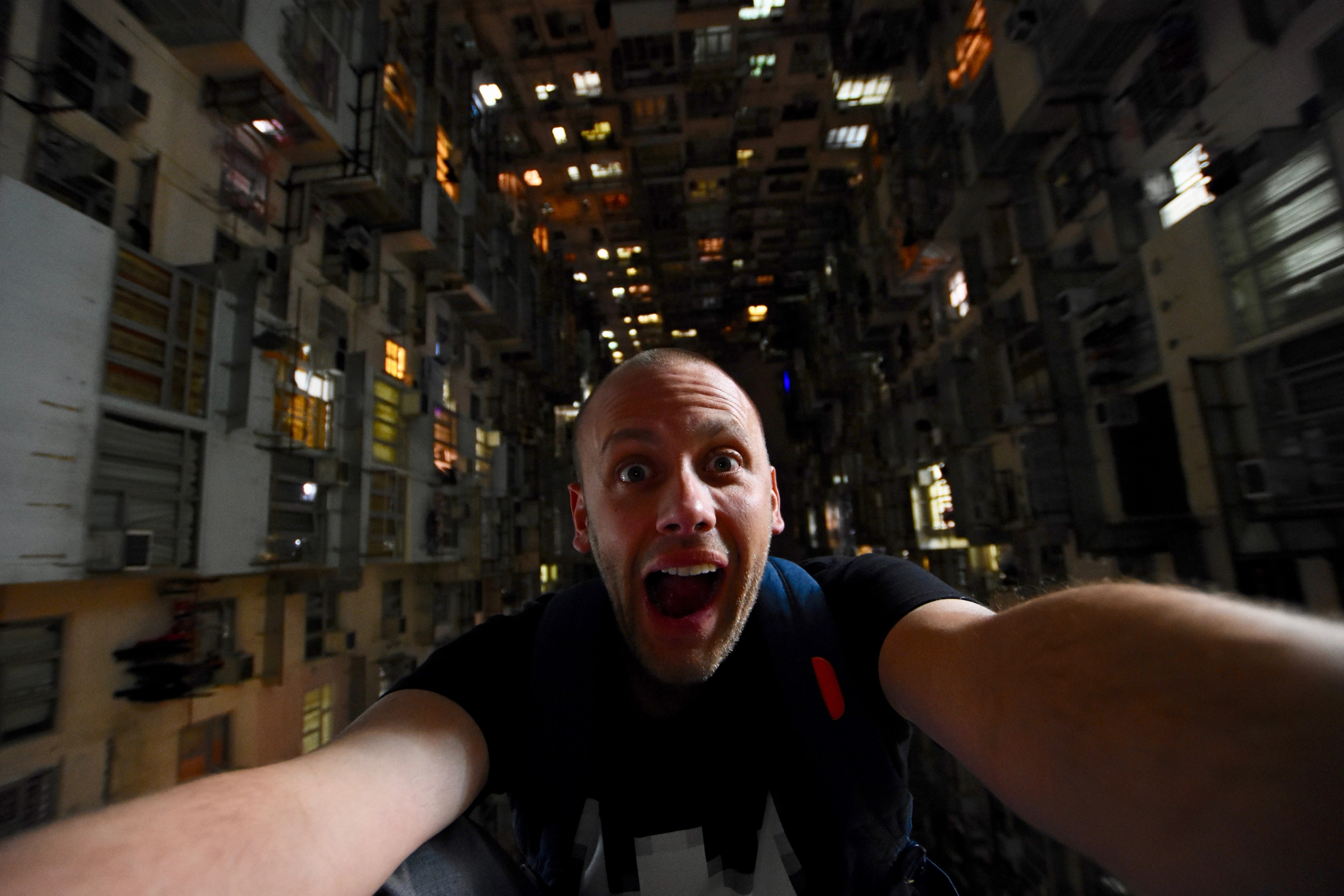 A man takes a close selfie with stacked apartment buildings behind him.