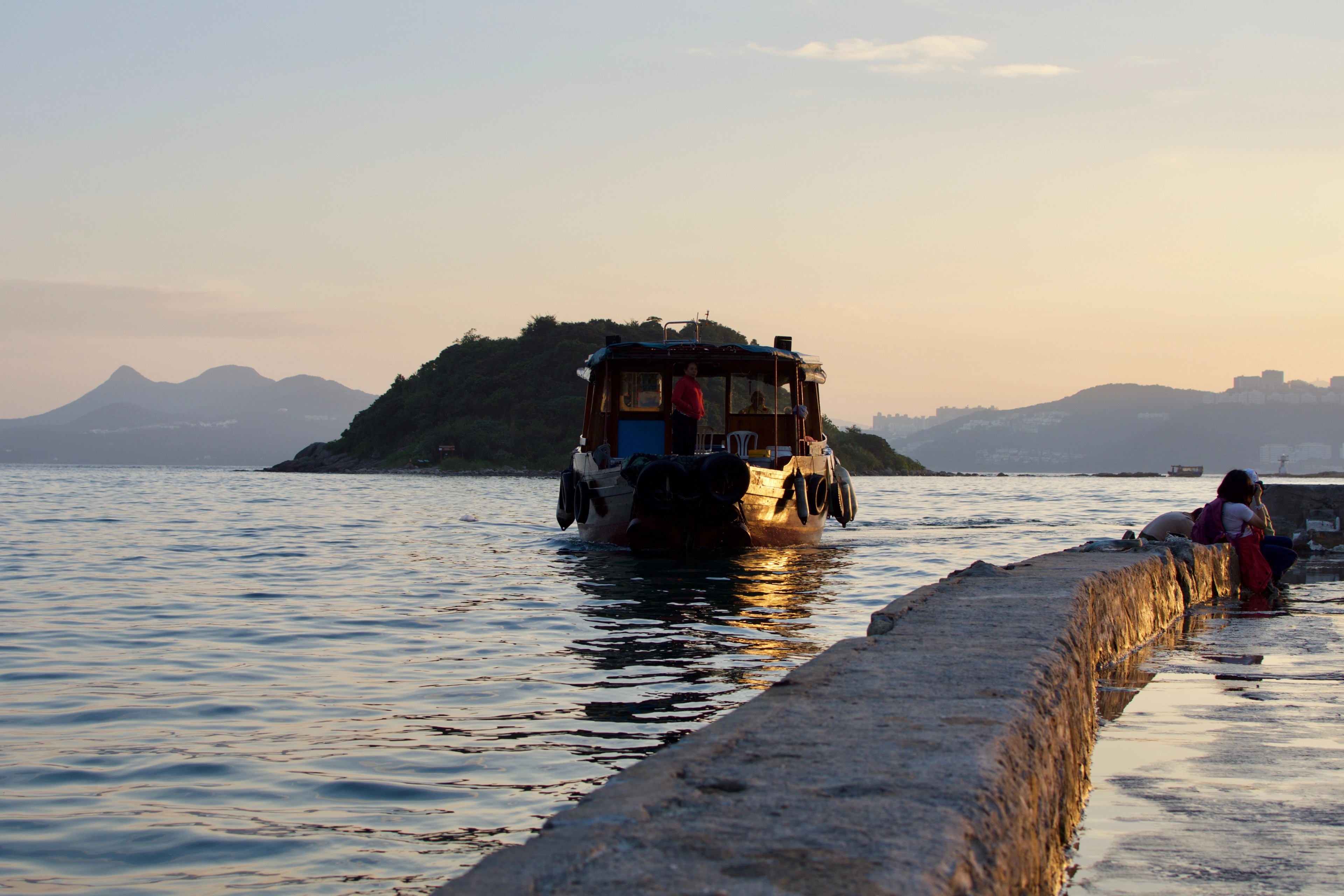 A small boat travels beside a concrete pier with islands in the distance.
