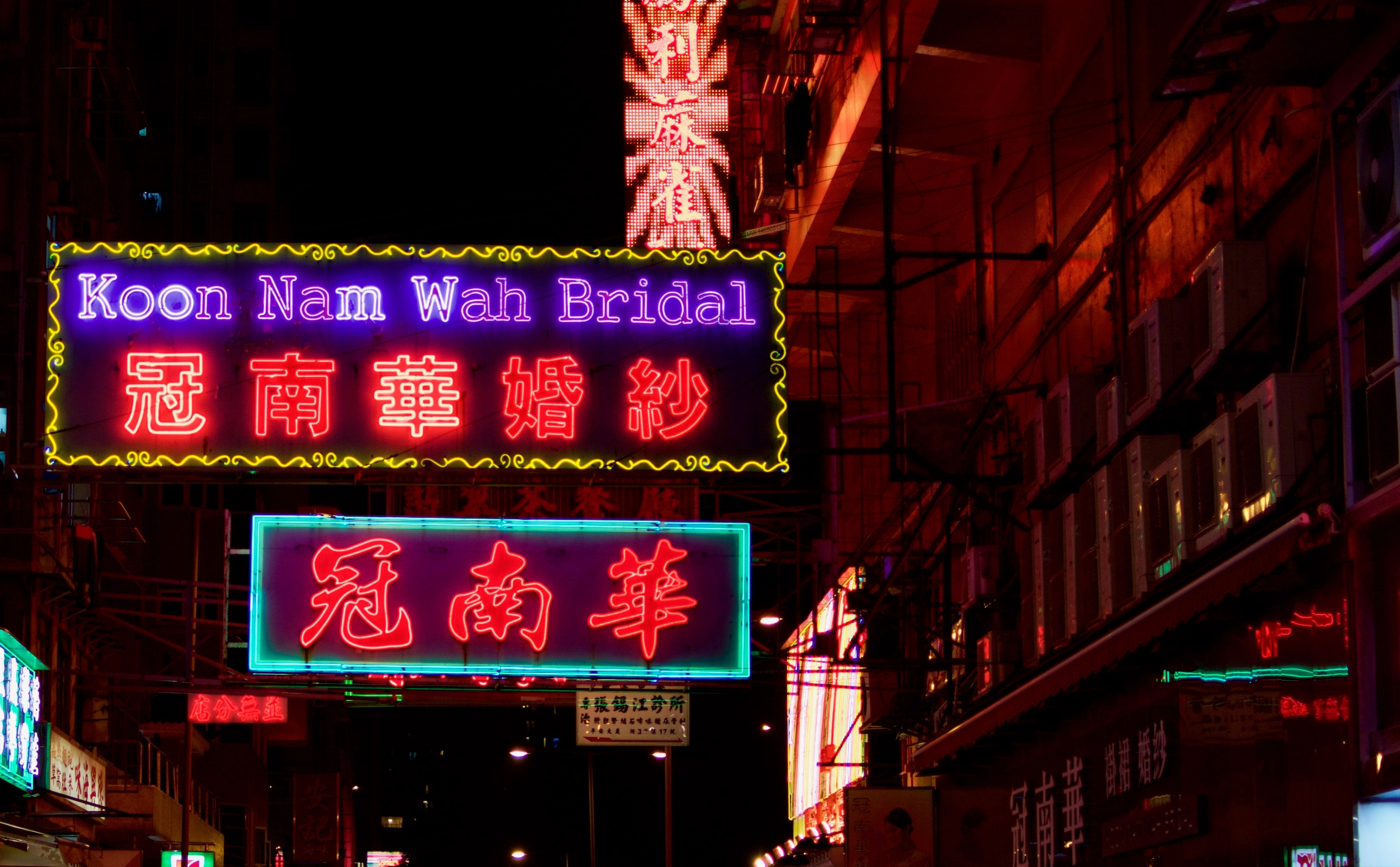Bright red, blue, and green neon shop signs fill a narrow night street.