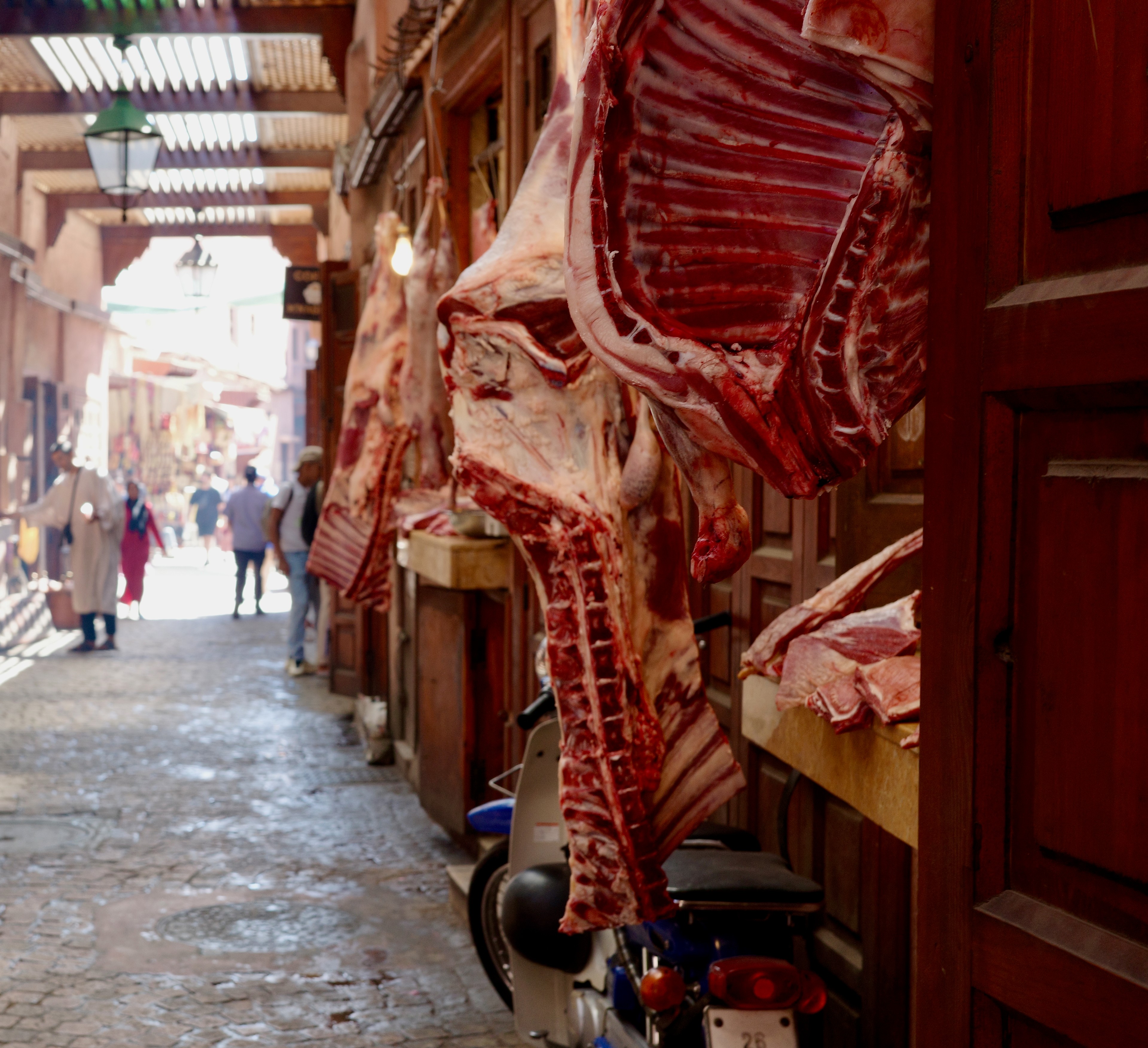 Cuts of meat hang outside a butcher shop on a narrow market street.