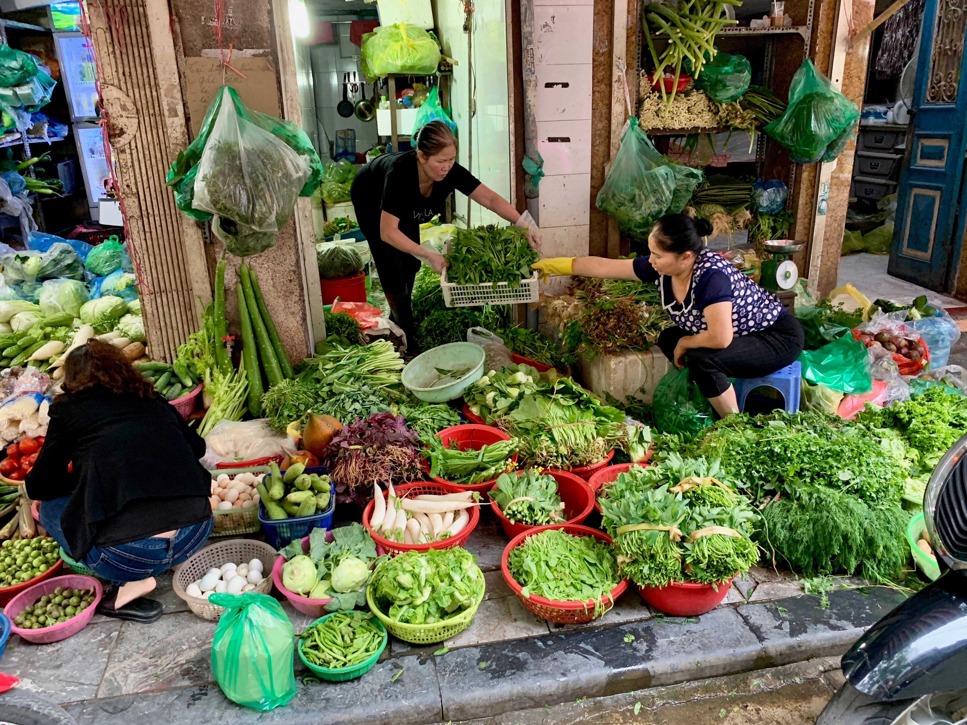 Customers gather around a market stall filled with leafy vegetables.