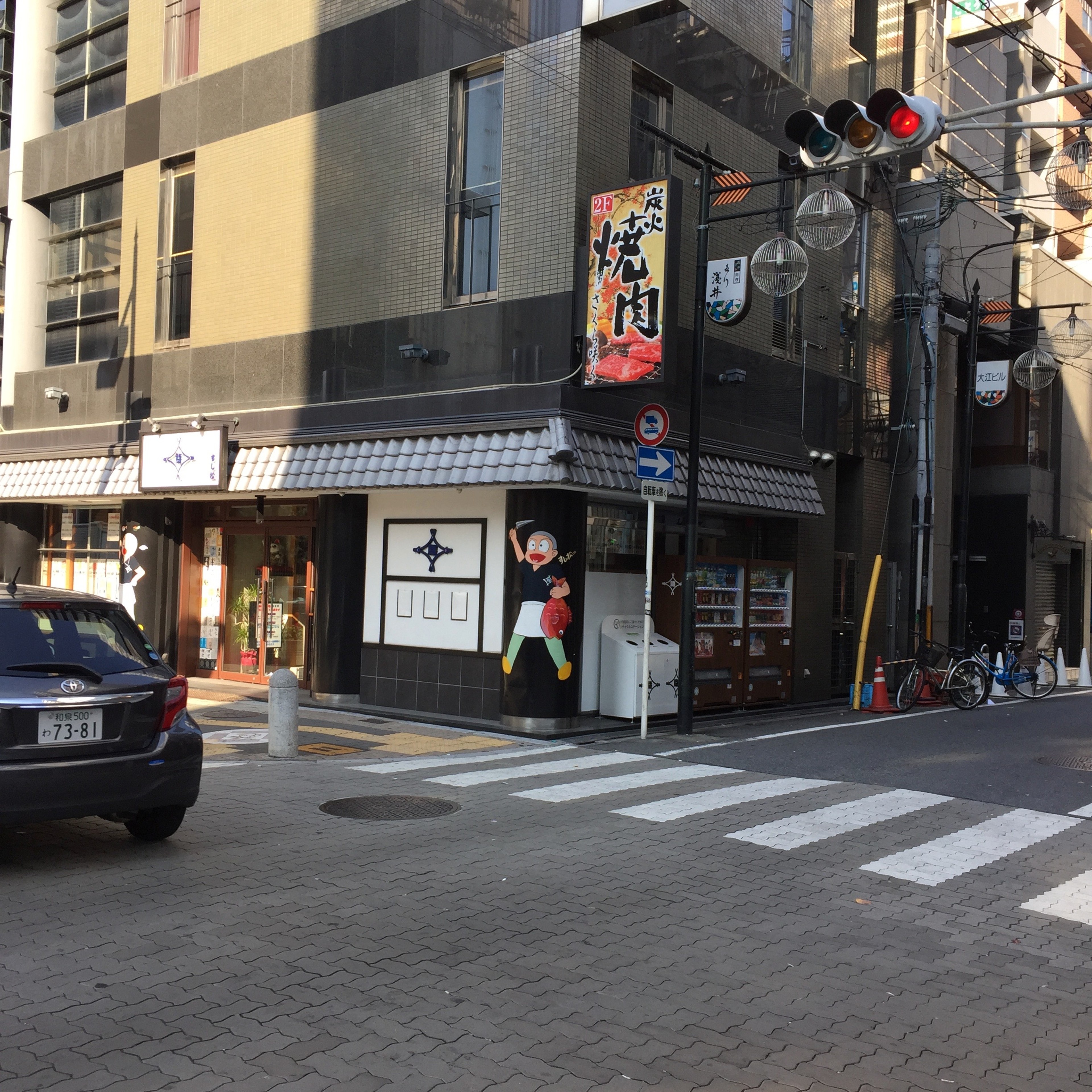 Pedestrians wait at a street corner beside storefronts and a crosswalk.