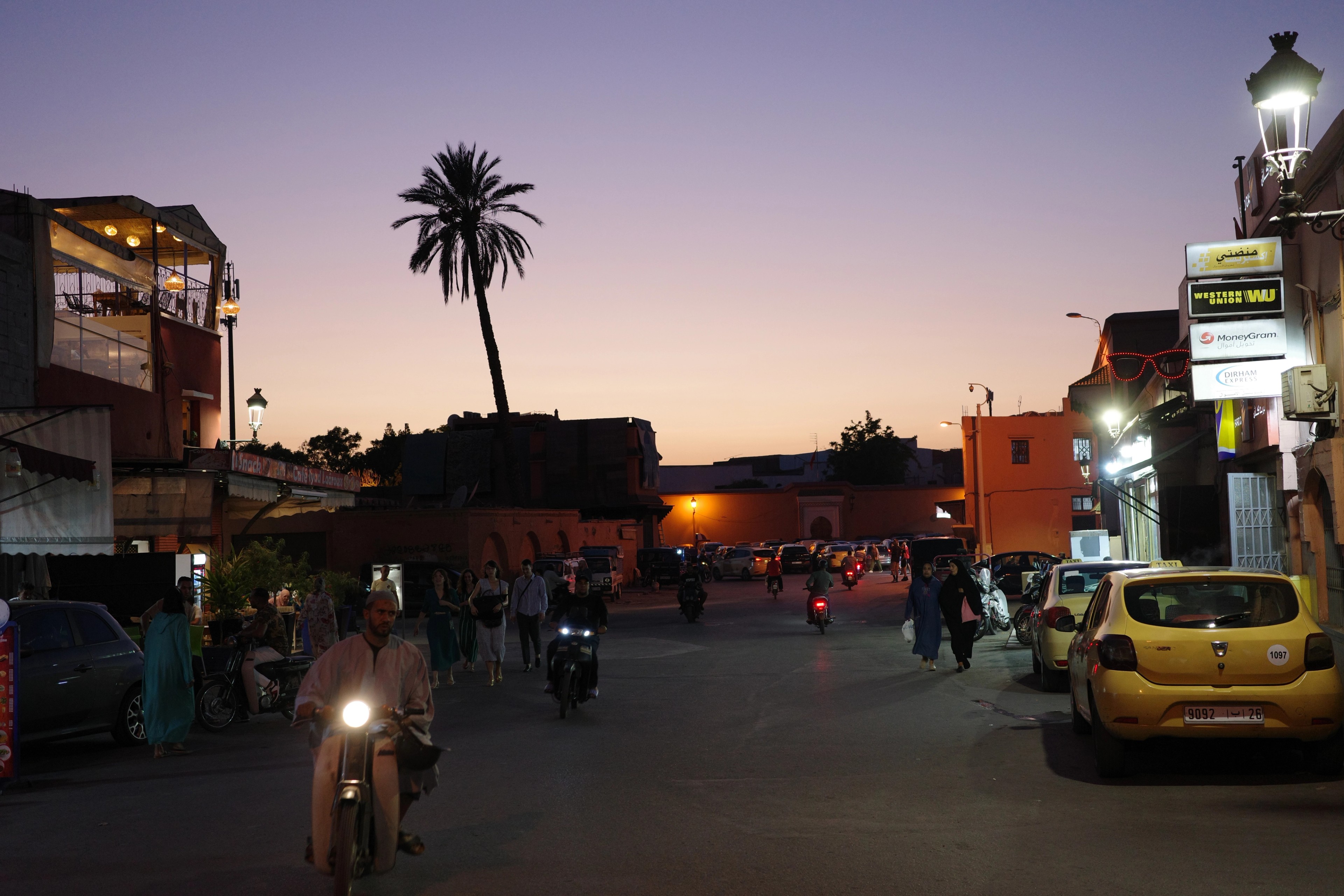 A busy square at dusk is lined with palm trees, vehicles, and market stalls.