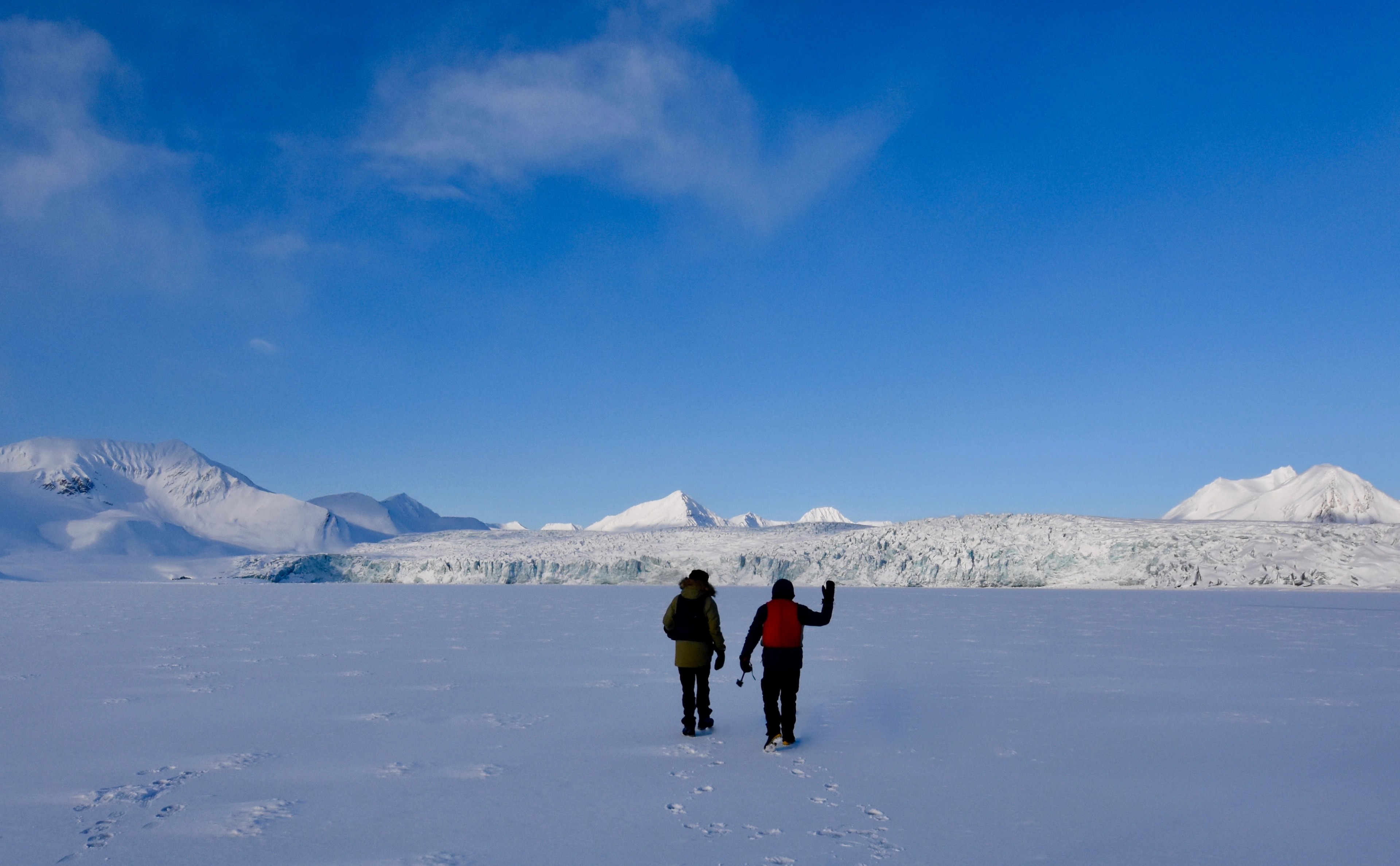 Two hikers cross an open snowfield toward distant mountains.
